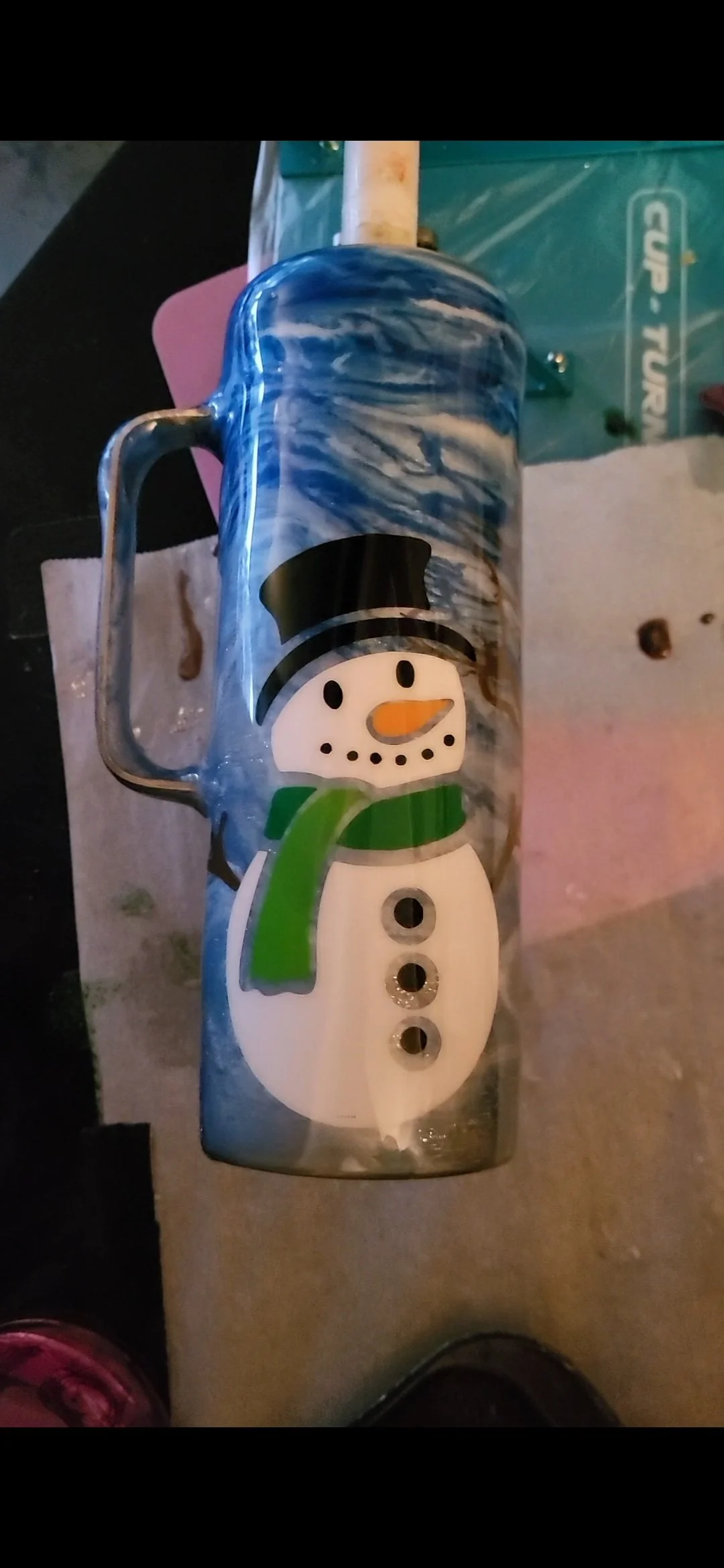 A glass mug with a snowman design, featuring a black top hat, orange carrot nose, green scarf, and buttons, on a surface with paper towels and a blue plastic bag nearby.