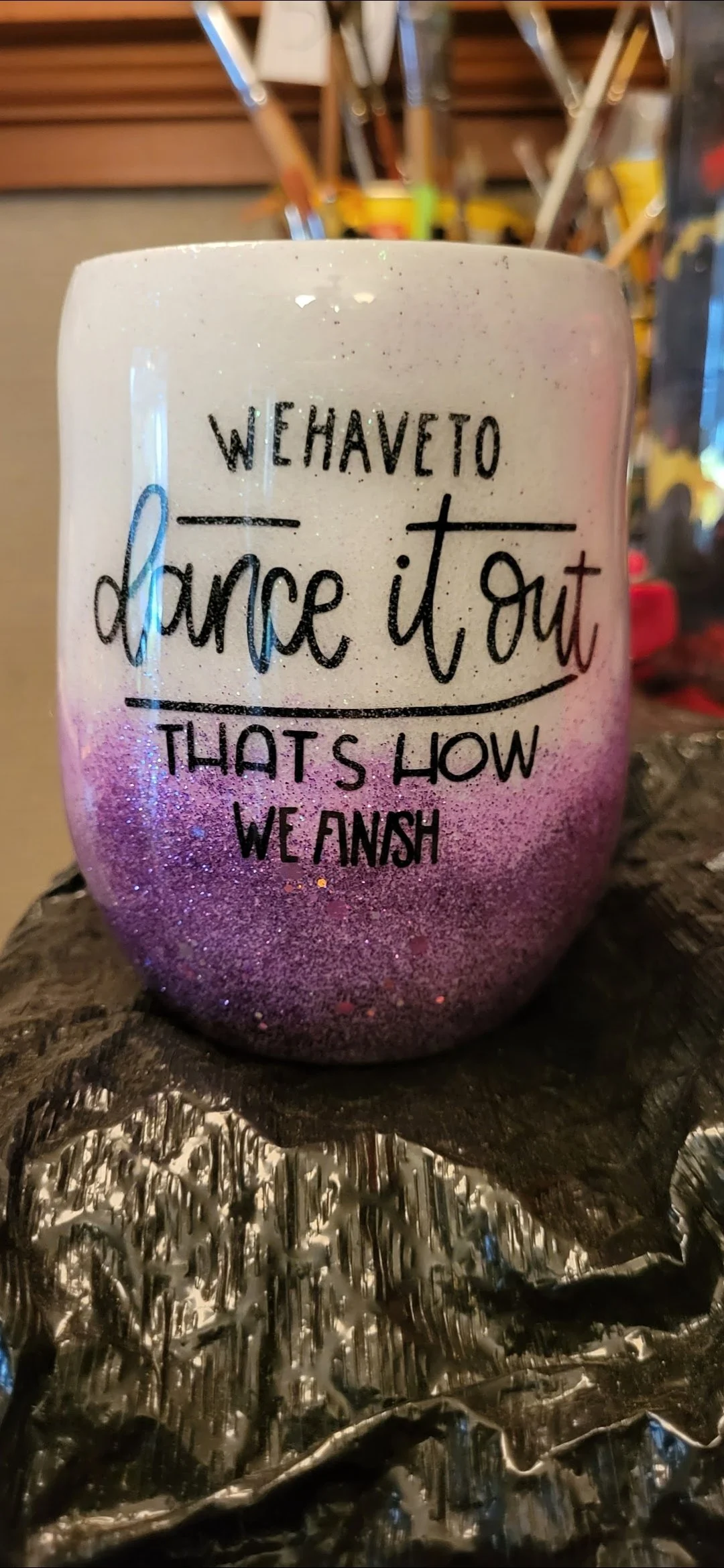 A decorated candleholder with a motivational quote in black text that says, 'We have to dance it out, that's how we flash.' The candleholder is pink and purple with glitter, placed on a wooden surface with a shiny metallic cloth underneath, and blurr