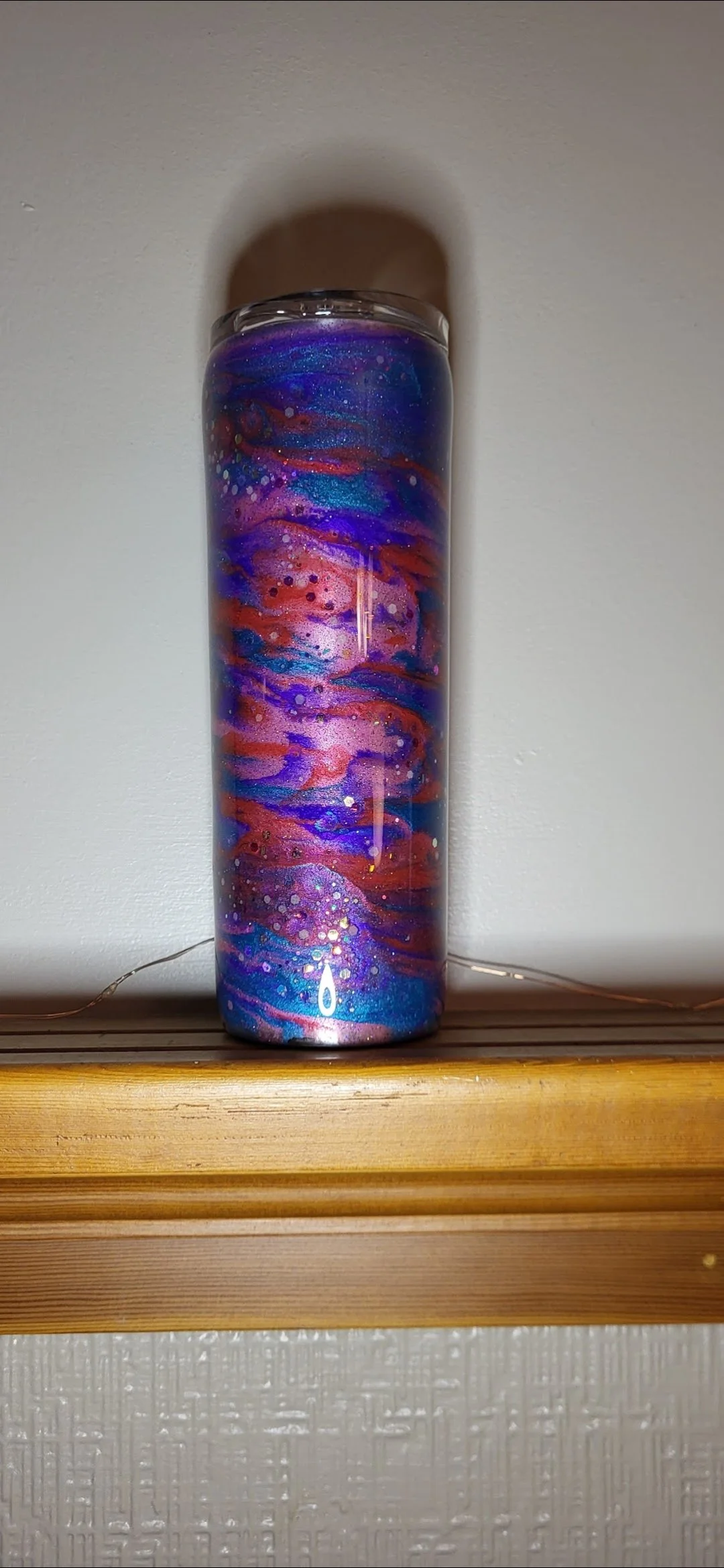 A tall, cylindrical glass jar with a galaxy-themed galaxy design featuring blue, purple, red, and pink swirls and specks of glitter, placed on a wooden surface against a plain wall.