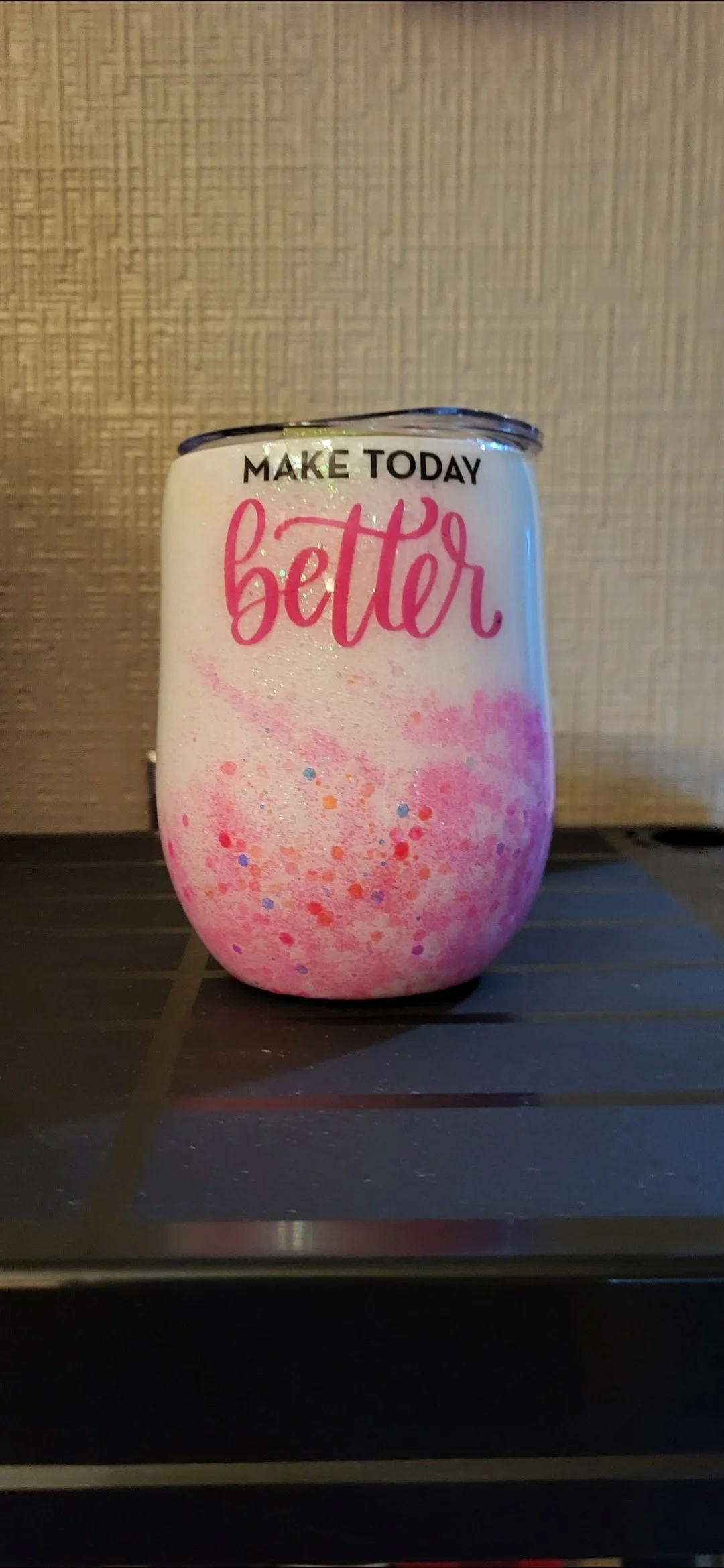 A decorated tumbler with pink, orange, purple, and blue splatters and the words "MAKE TODAY better" written on it, placed on a dark surface against a textured off-white background.