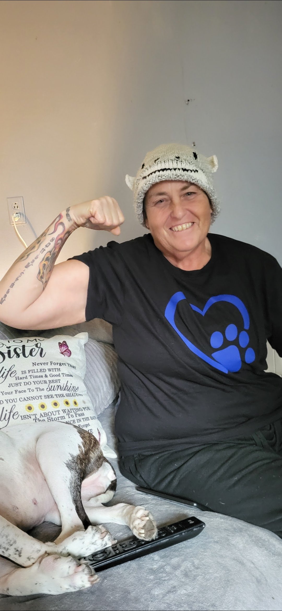 A woman in a black t-shirt with a blue heart and paw print design, wearing a knit hat, smiling and flexing her bicep. She is sitting on a couch beside a dog with brown and white fur, which is lying with its paws on a remote control. There is a decora