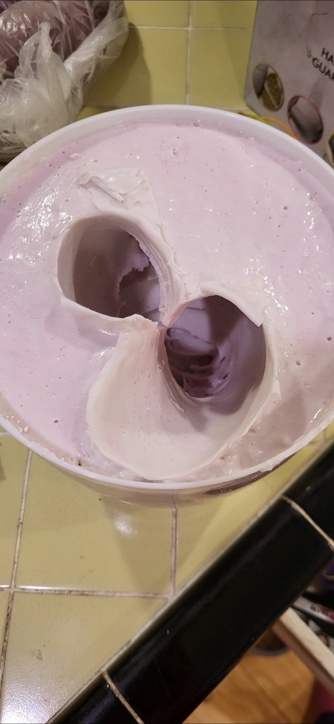 A pale pink tub of ice cream with a large scoop taken out, revealing the creamy texture inside, placed on a tiled floor.