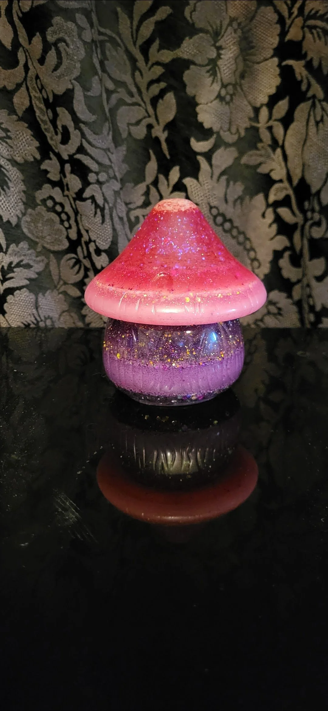 A glass mushroom-shaped decorative object with a pink and purple color scheme, glitter and sparkles, placed on a reflective black surface with a patterned wallpaper background.