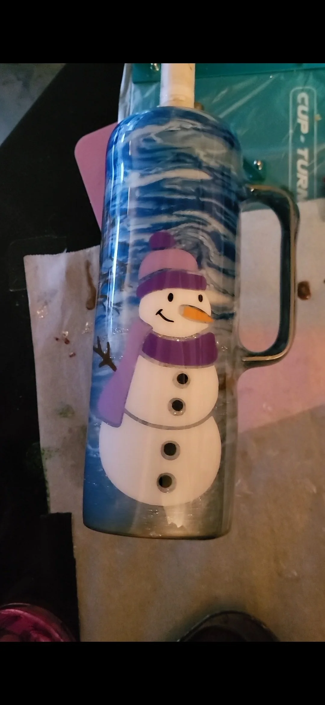 A snowman-themed mug with a smiling snowman wearing a purple hat and scarf, with a carrot nose, black buttons, and a cheerful expression, against a blue swirling background.