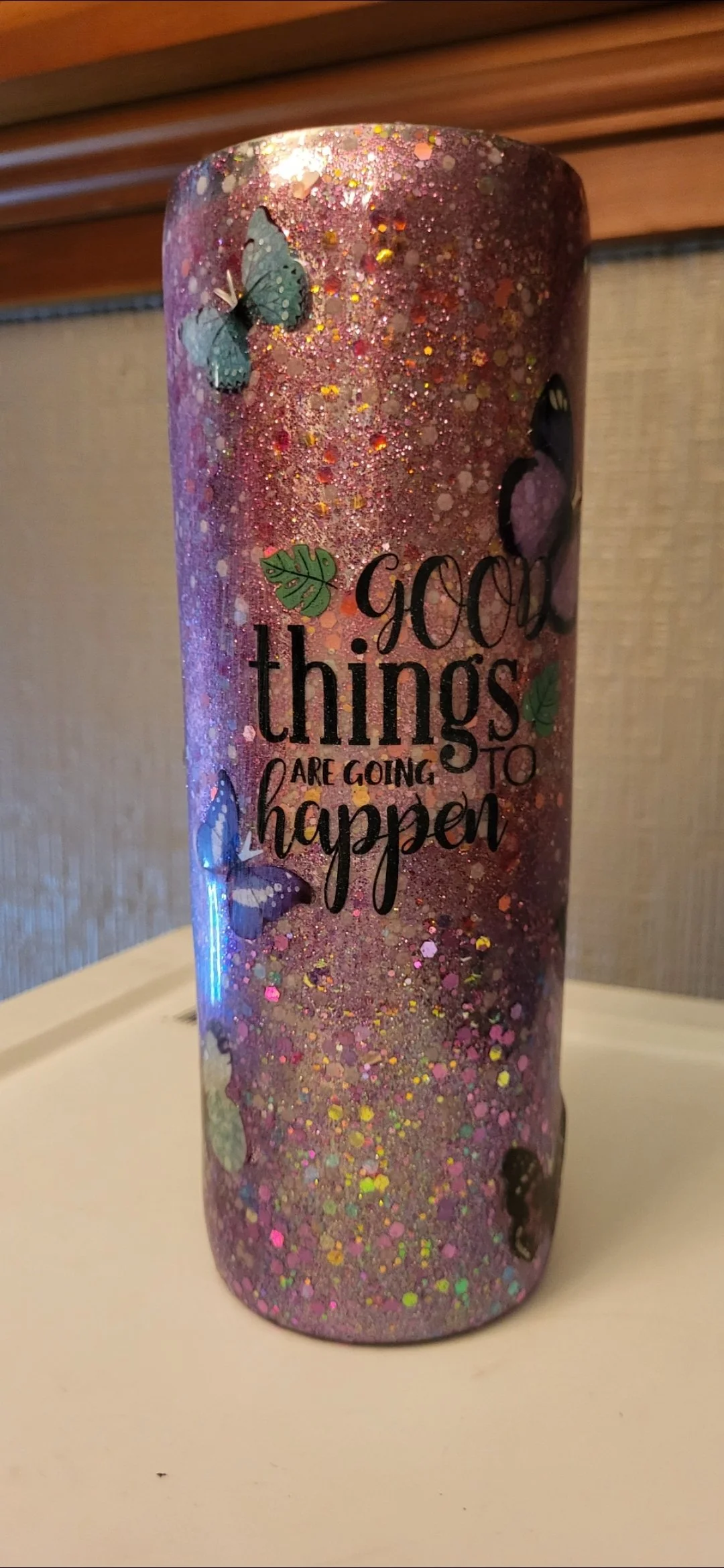 A tall, pink glitter candle decorated with colorful butterflies and leaves, with the motivational quote 'Good things are going to happen' in black and pink lettering.