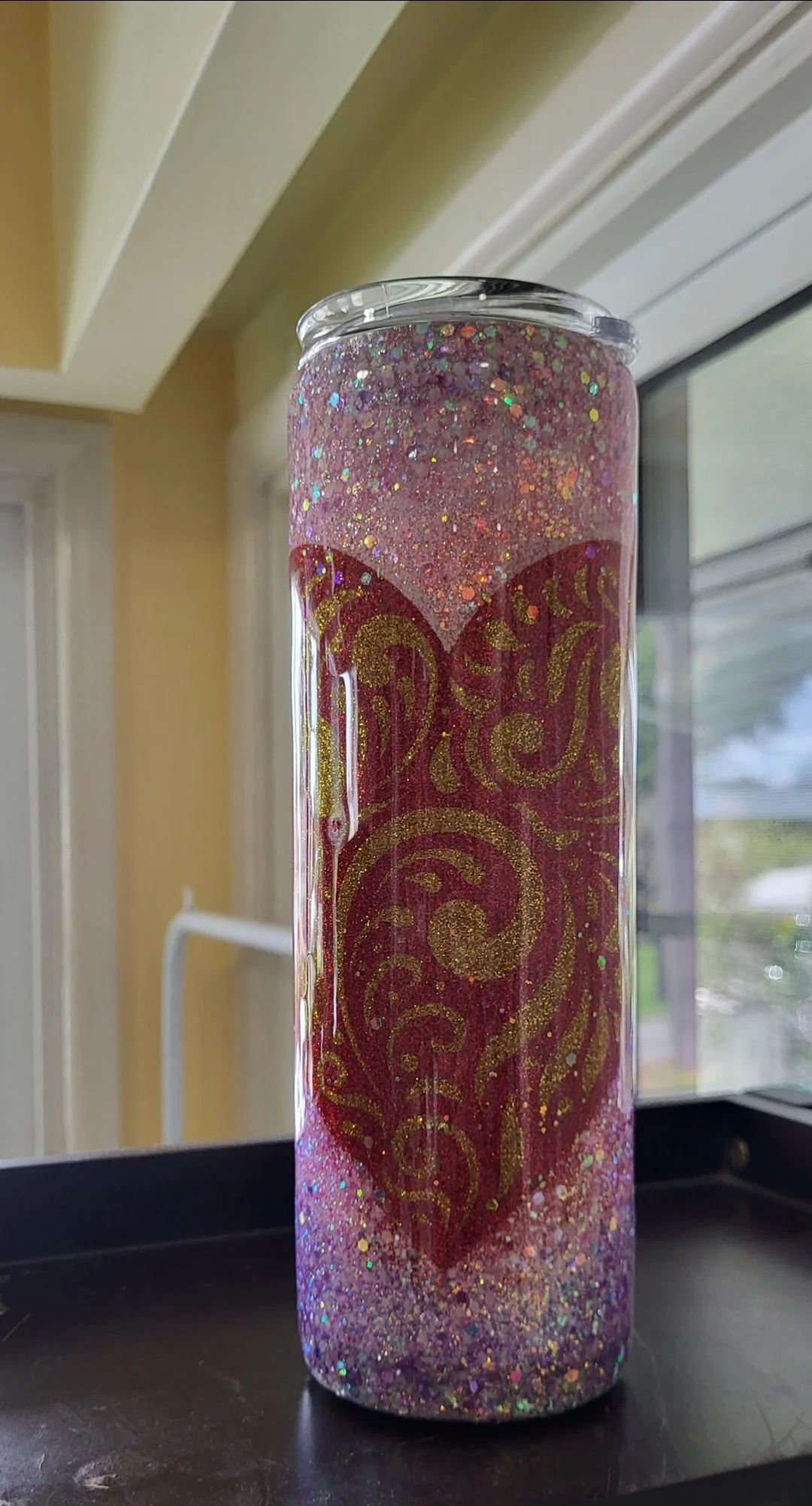 A tall tumbler with glitter and gold heart design, with a pinkish-purple glittery liquid inside, placed near a window.
