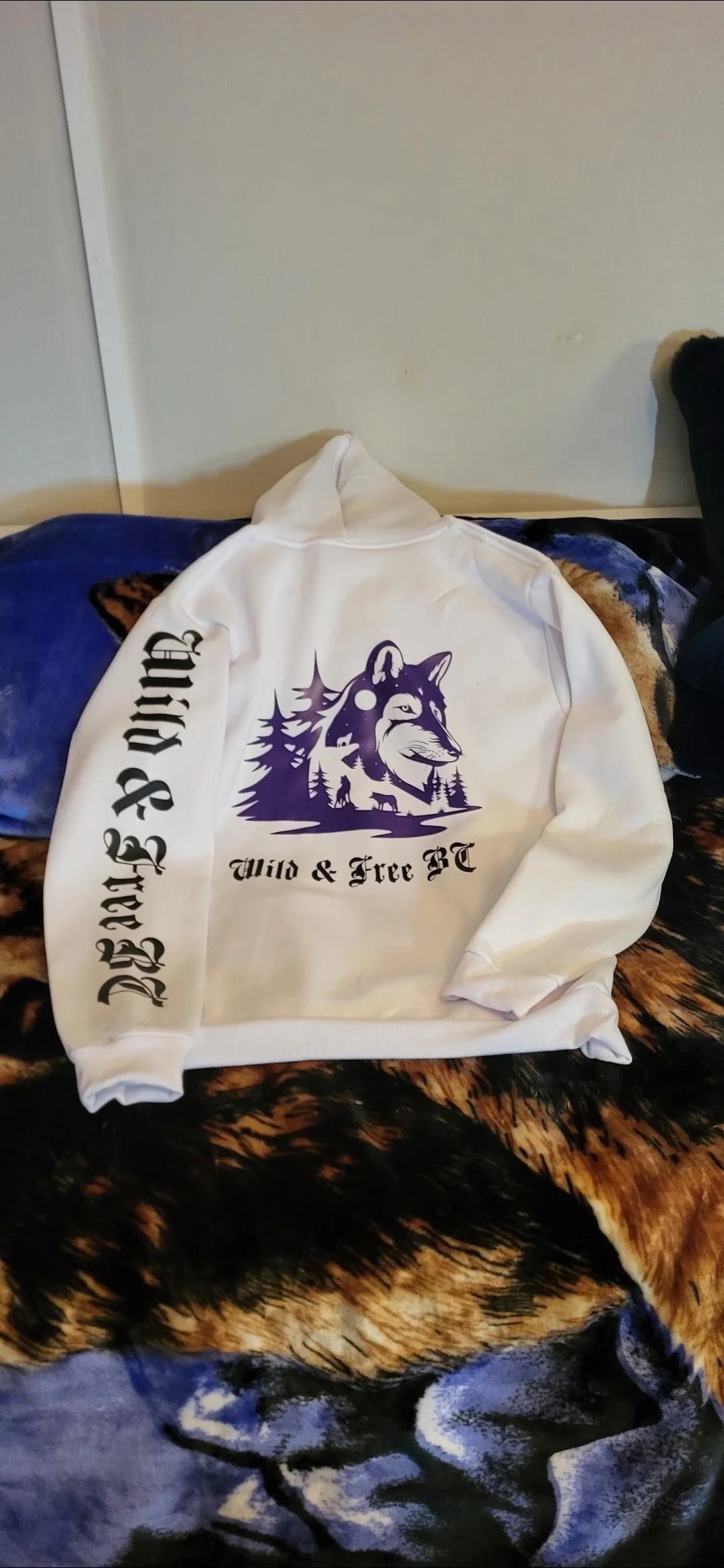White hoodie with a purple graphic of a wolf and trees, and black text that reads 'Wild & Free BC' on the front. The hoodie is laying on a bed with a black and brown animal print blanket and a blue pillow.