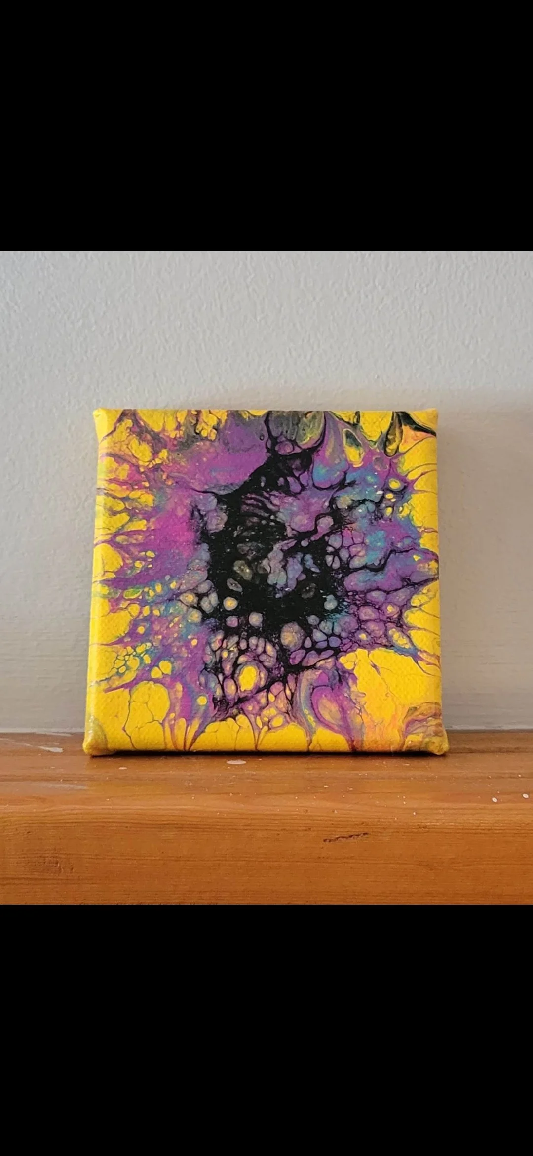 Small abstract painting with black, purple, yellow, and blue splatters on a yellow background, leaning against a light-colored wall.