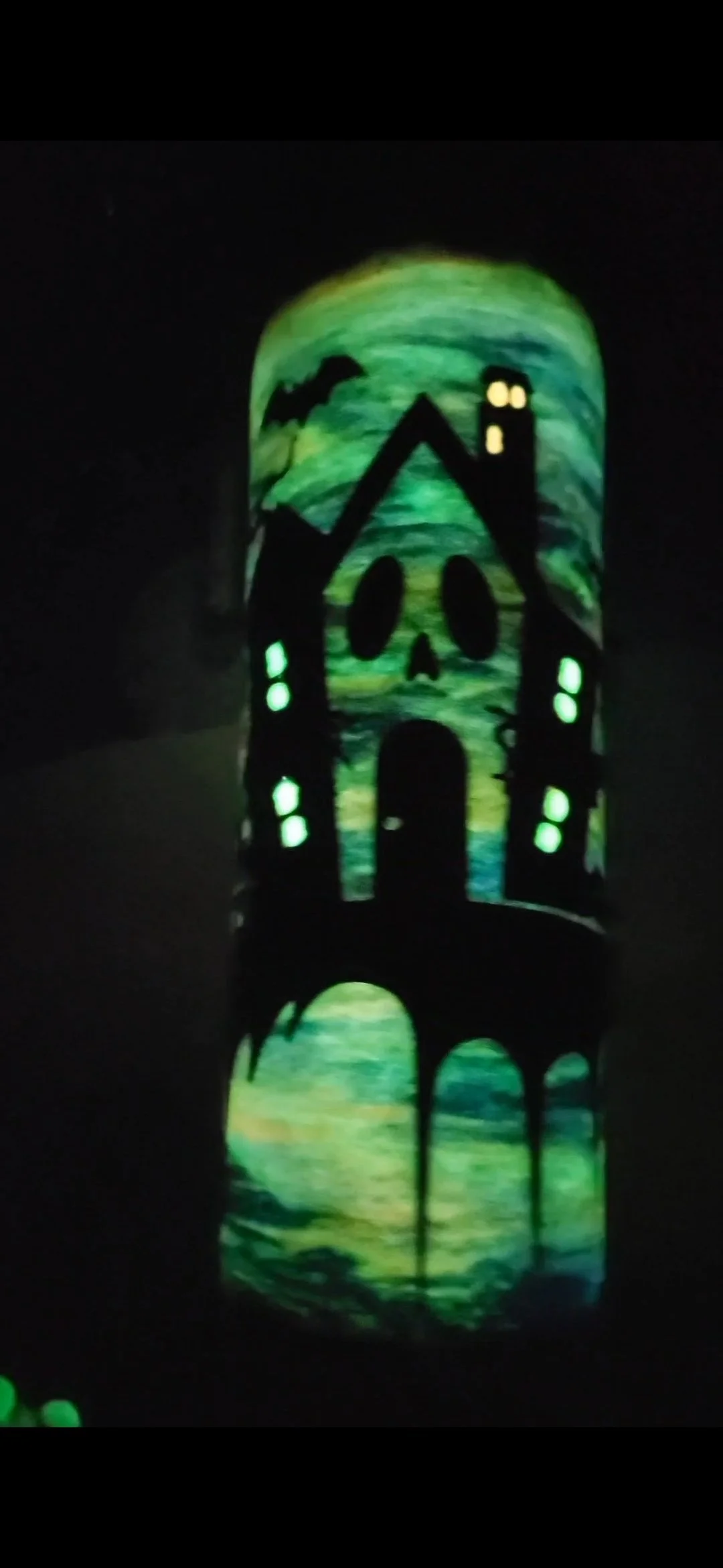 Glow-in-the-dark Halloween decoration of a haunted house with a skull face, glowing windows, a bat, and a full moon, set against a dark background.