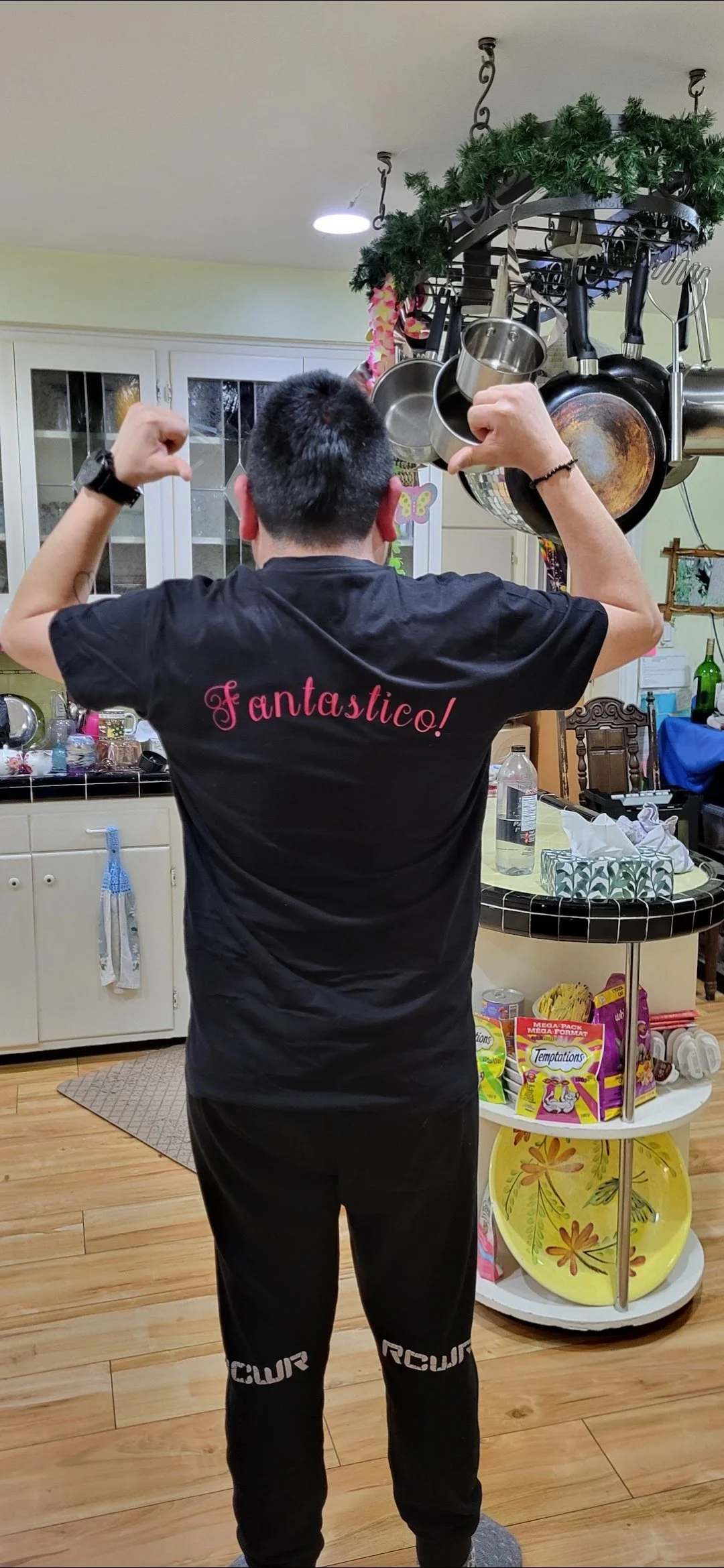 A young man standing in a kitchen, facing away from the camera, flexing his muscles. He is wearing a black t-shirt with 'Fantastico!' written on the back in pink, and black pants. The kitchen has a hanging pot rack with pots and pans, a small white r