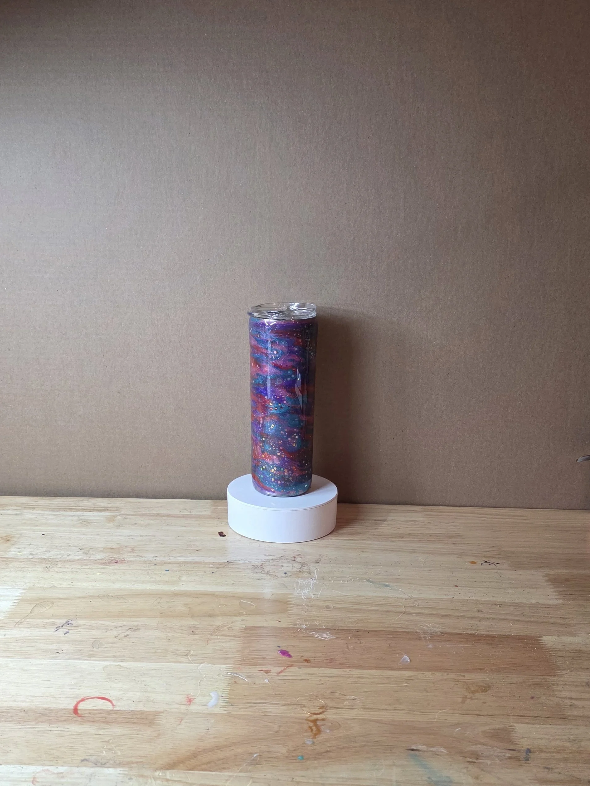 A colorful galaxy-themed candle or object on a white round stand, placed on a wooden table with paint stains, against a plain beige wall.