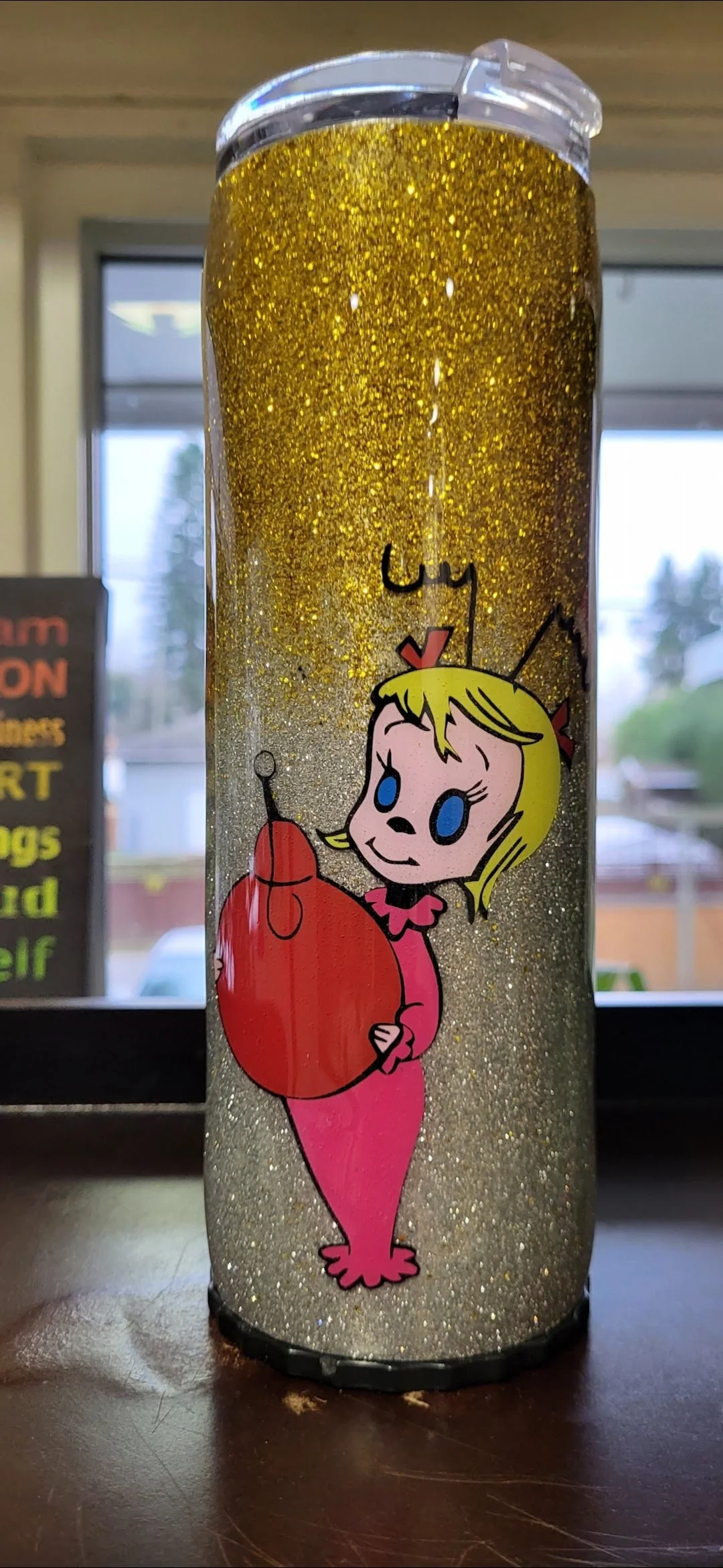 A tumbler with gold glitter and an illustration of a cartoon girl with blonde hair, blue eyes, wearing a pink dress, holding a red bomb with a lit fuse.