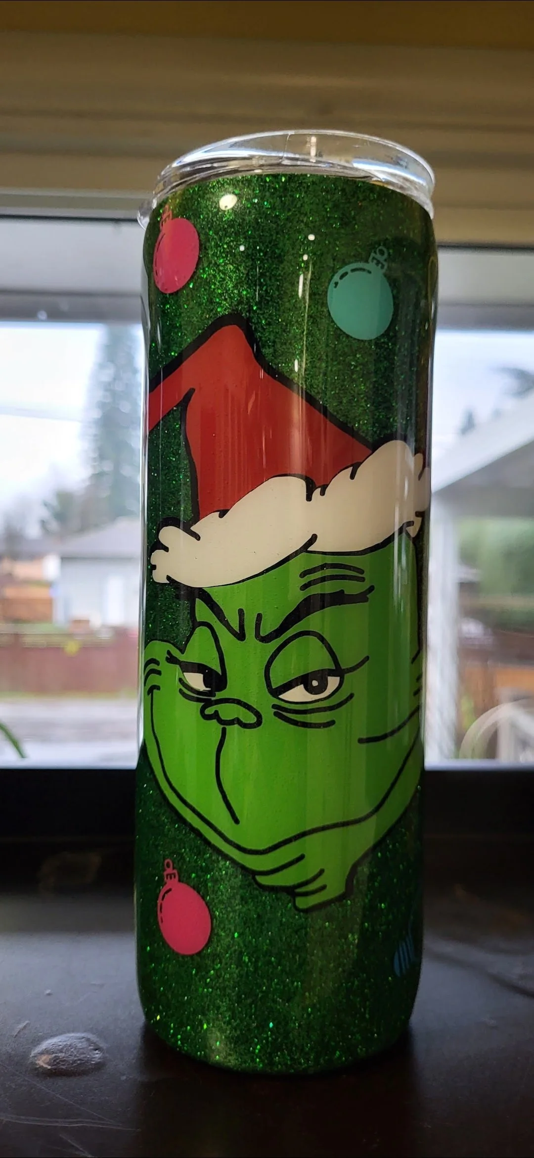 A bottle with a green glittery background, featuring a cartoon Grinch face wearing a Santa hat, and small pink and blue decorative circles.