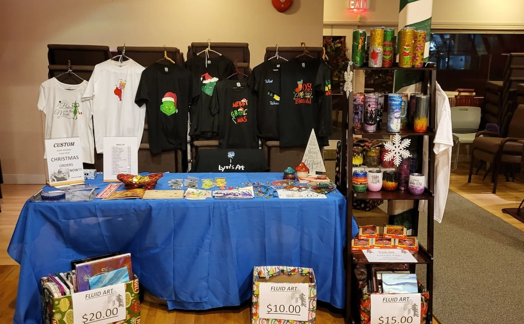 A holiday craft booth with Christmas-themed shirts hanging on a wall in the background, a table with various small art items, and a shelving unit filled with colorful painted jars and candles. Signage indicates prices for liquid art items. Decoration
