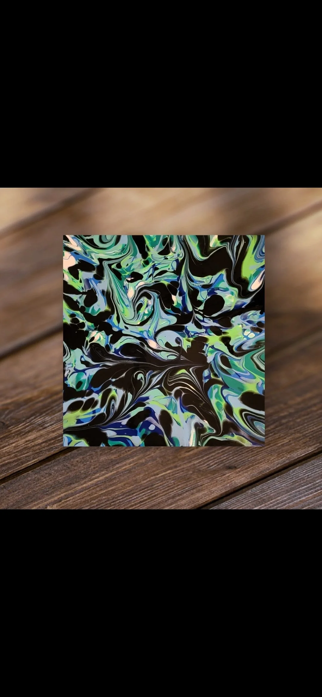 Abstract swirl painting with green, blue, black, and white colors on a square canvas placed on a wooden surface.