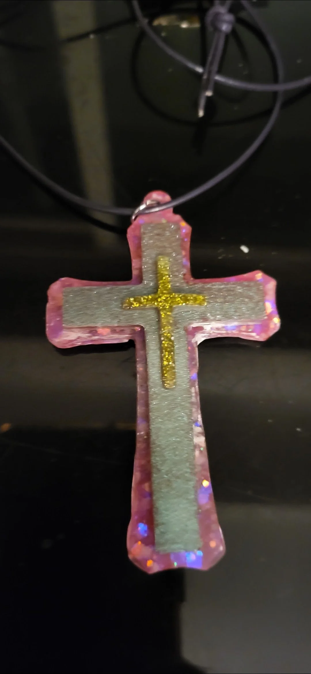 Pink and silver cross-shaped ornament with gold glitter cross in the center, hanging from a black string.