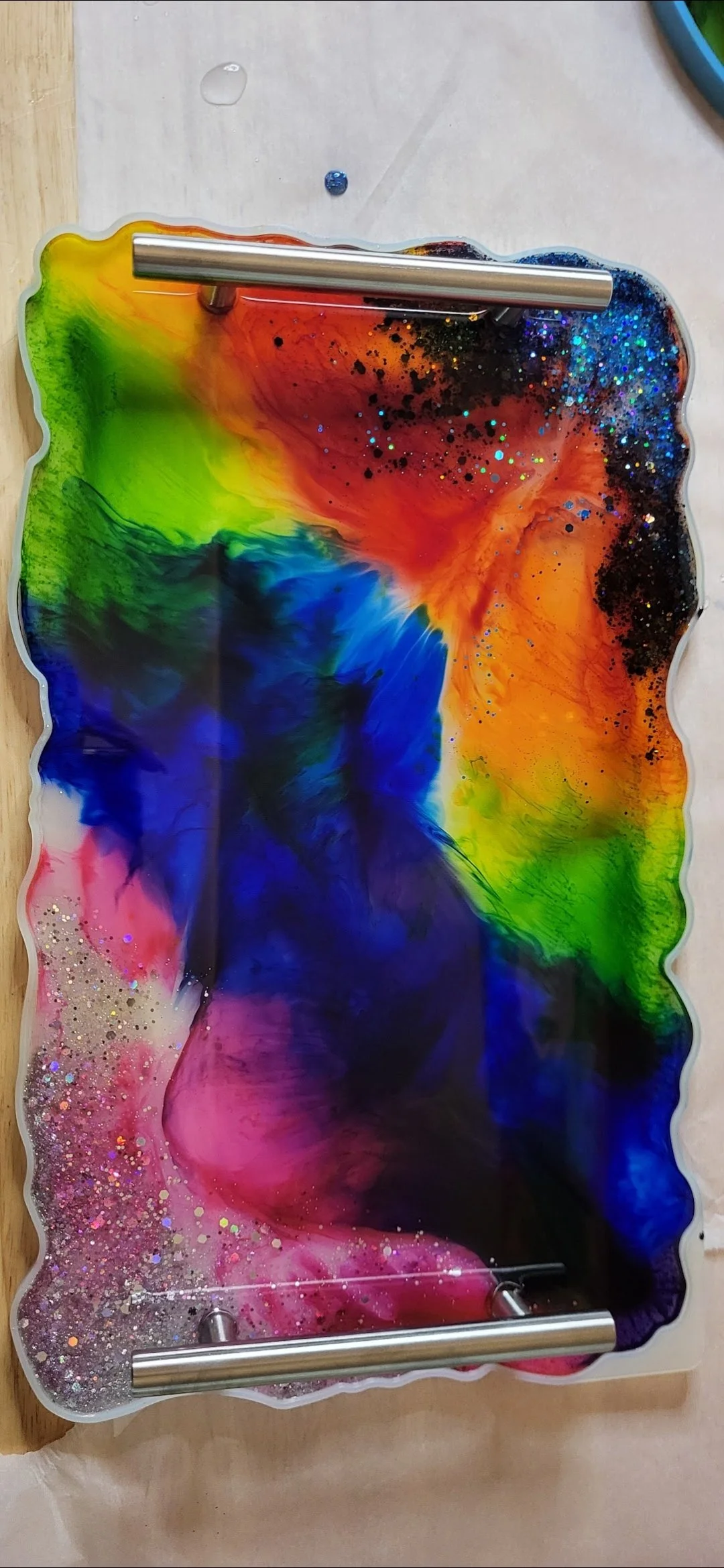 Colorful abstract resin art with swirling rainbow colors, glitter, and black speckles on a decorative tray with metal handles.