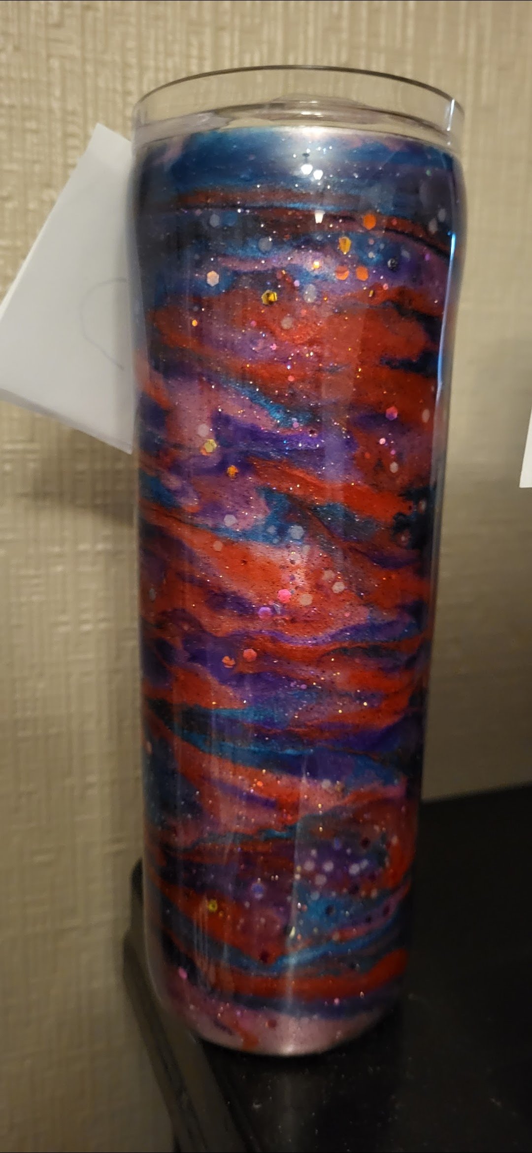 A tall cylindrical glass vase with a galaxy-inspired abstract design in red, purple, blue, and black colors with glitter accents, standing on a dark surface against a textured beige wall.