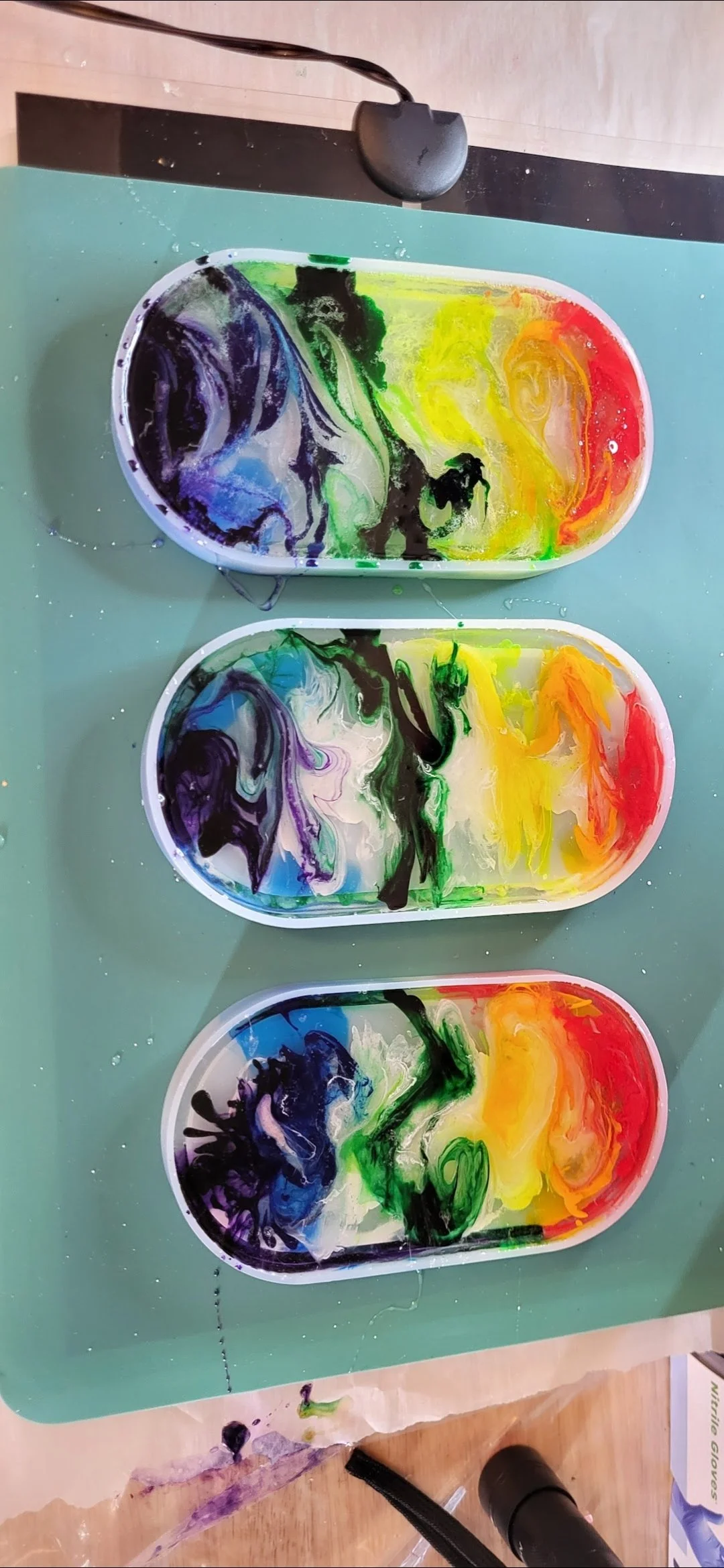 Three oval paint palettes with colorful swirling paints, placed on a green surface. The palettes contain a mix of black, purple, blue, green, yellow, orange, and red paints, with some paint splatters and spills around them.