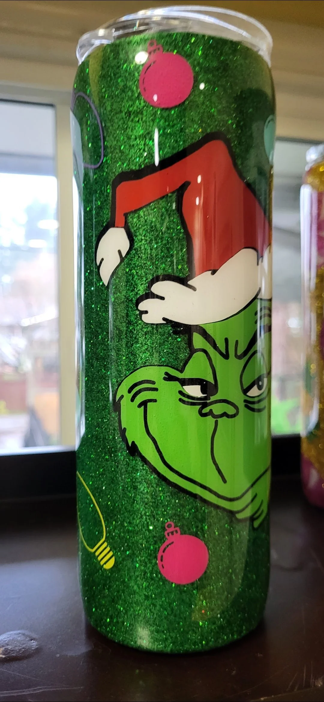 A glittery green tumbler decorated with the Grinch character from Dr. Seuss, featuring the Grinch's face with a sly smile and half-closed eyes, and the top part shows the Grinch's head and hand holding a red ornament while the bottom features a Chris