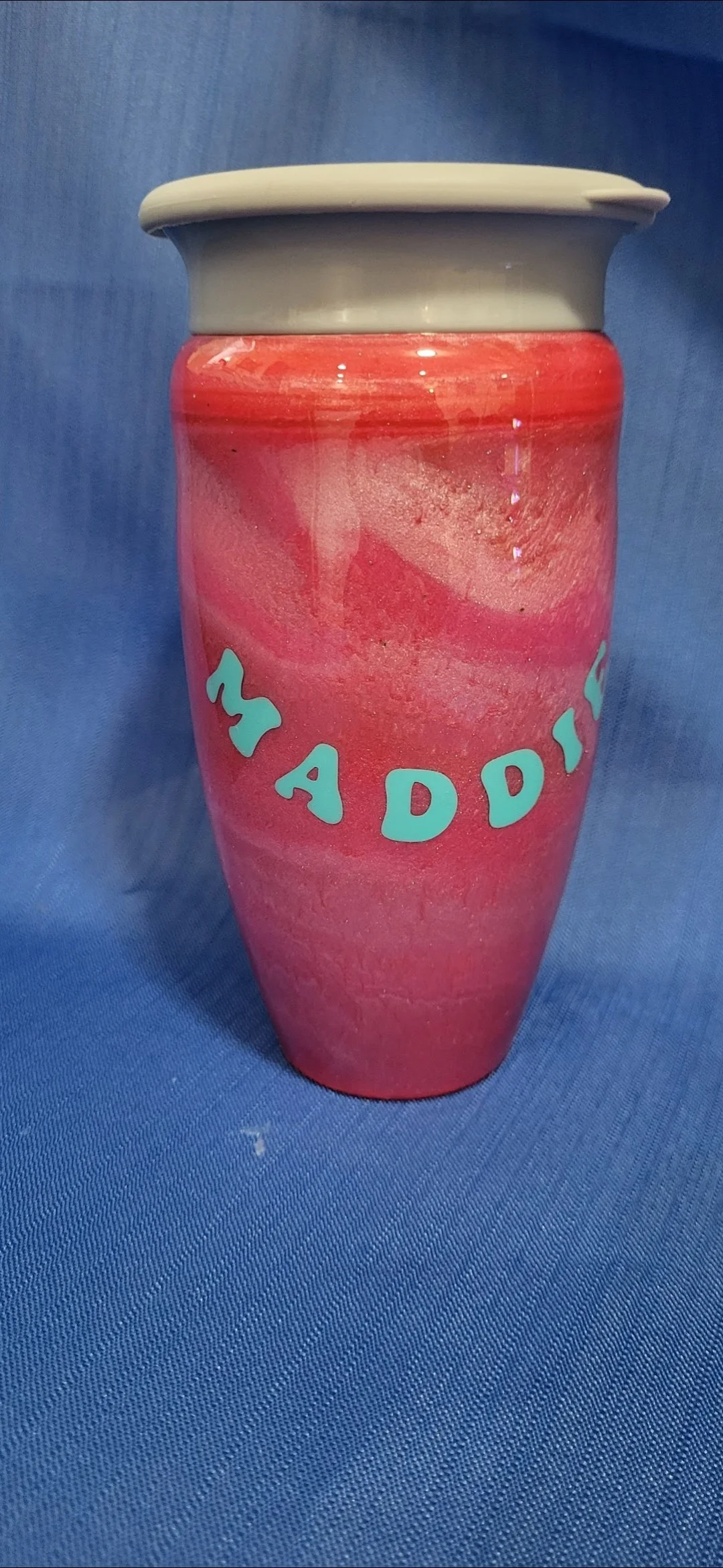 A pink and red painted mug with the words 'MY ADDI...' written on it in light blue letters, set on a blue fabric background.