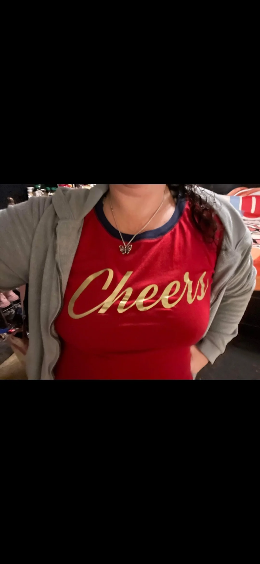 A woman wearing a red t-shirt with the word 'Cheers' printed in gold, paired with a gray zip-up hoodie and a butterfly-shaped necklace.