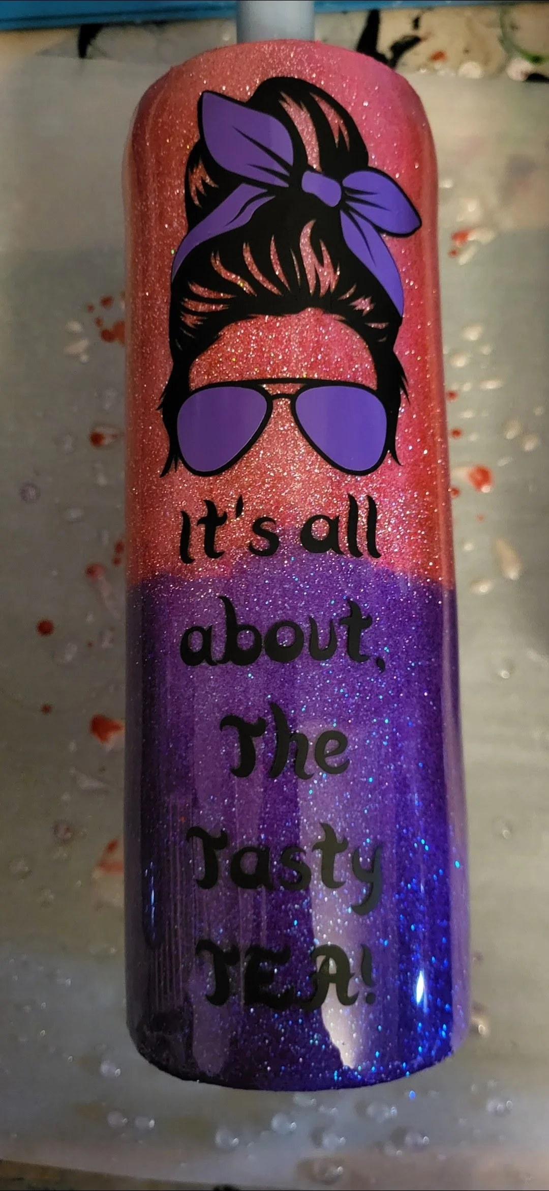 A pink and purple glittery tumbler with an illustration of a woman's hair tied with a purple bandana, wearing purple sunglasses and the text 'It's all about. The Tasty JEA!'