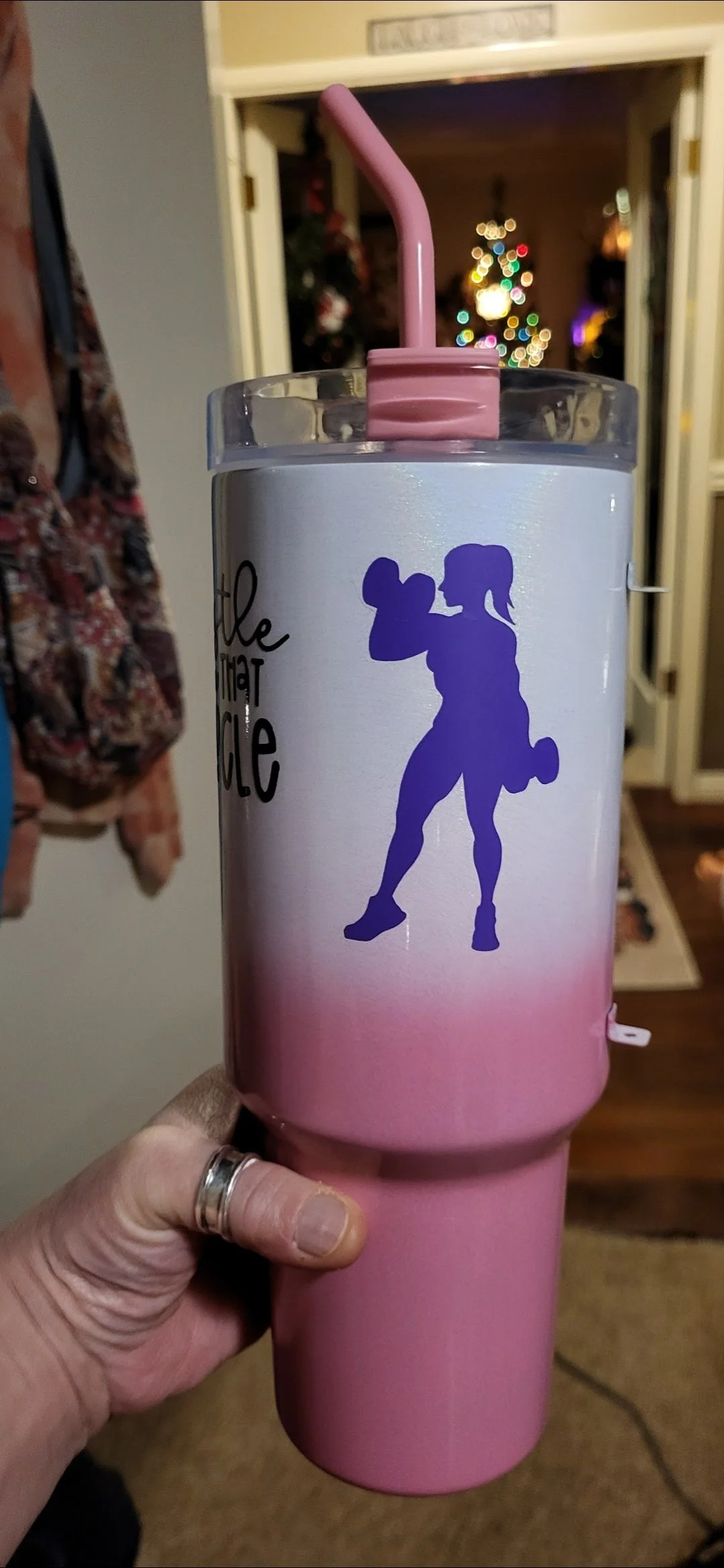 Pink tumbler with a silhouette of a woman lifting a dumbbell, set against a white background with pink gradient at the bottom, held by a person's hand with a ring. In the background, a Christmas tree with colorful lights is visible.