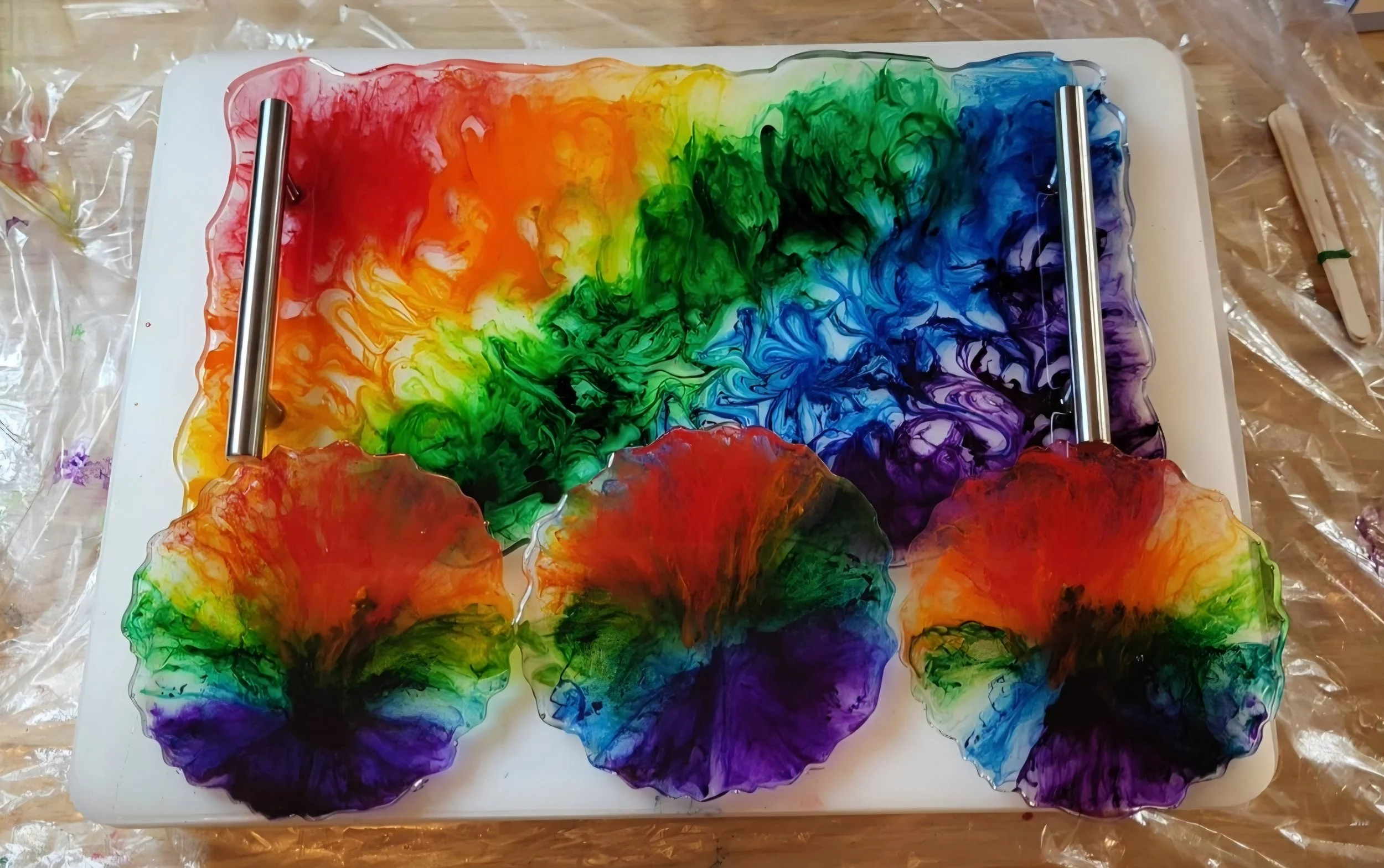 Colorful ink swirling in water, with three stained glass-like rainbow-colored flowers in front, set on a white rectangular surface.