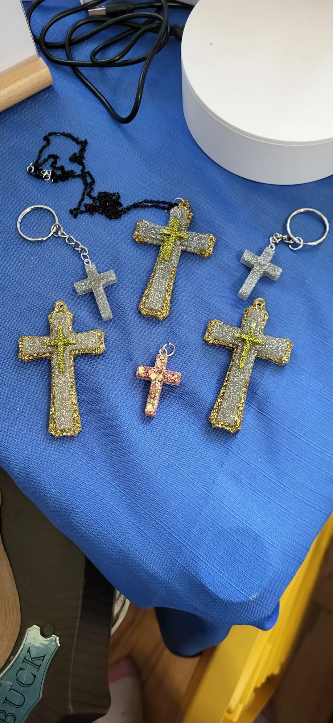 Various cross-shaped pendants and keychains with gold, silver, and pink glitter, laid out on a blue fabric surface.