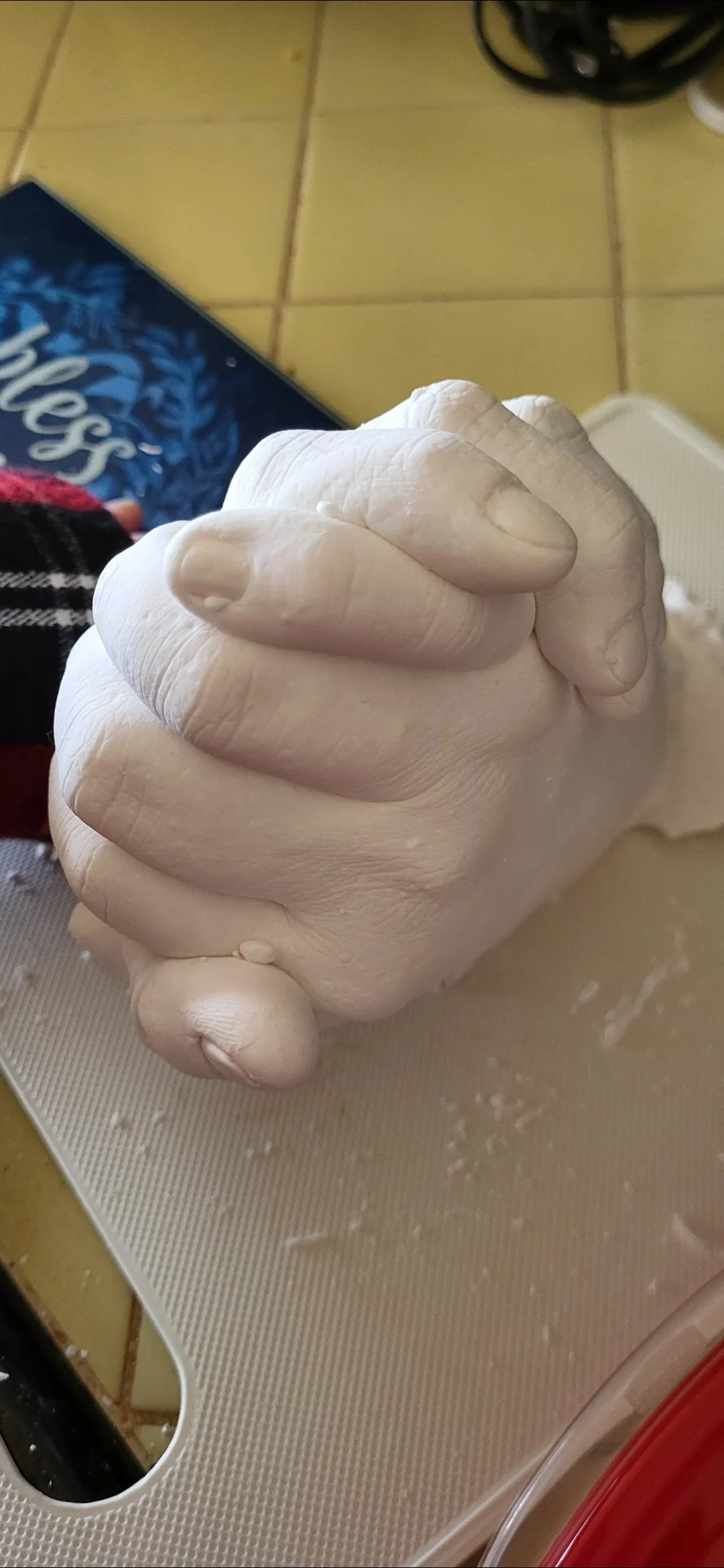 A close-up of a carved white sculpture of two clasped hands resting on a textured surface, with a blue box and tiled floor in the background.