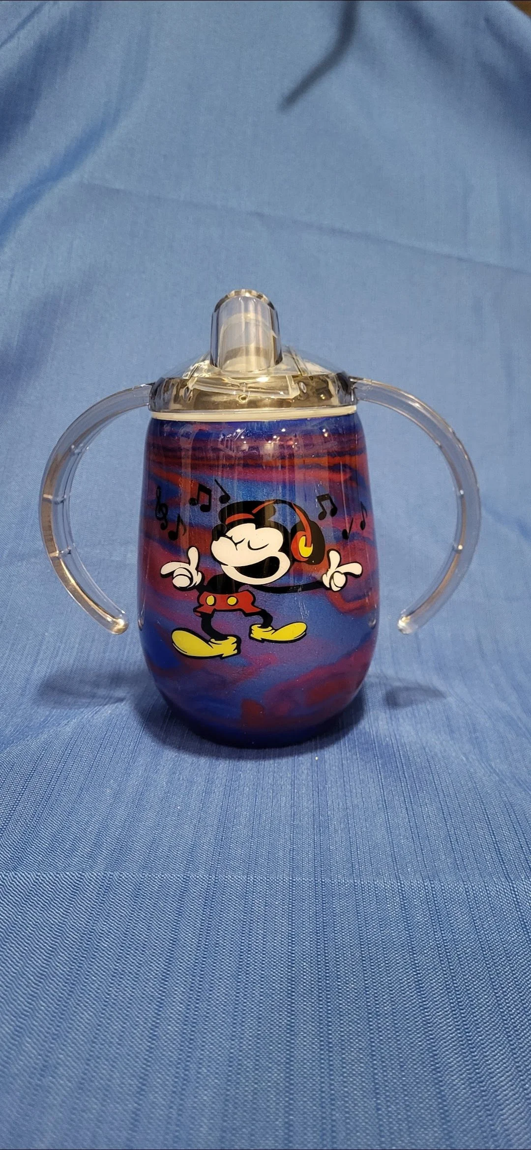 A baby sippy cup with a Mickey Mouse design, featuring Mickey wearing headphones and musical notes around him, placed on a blue fabric background.