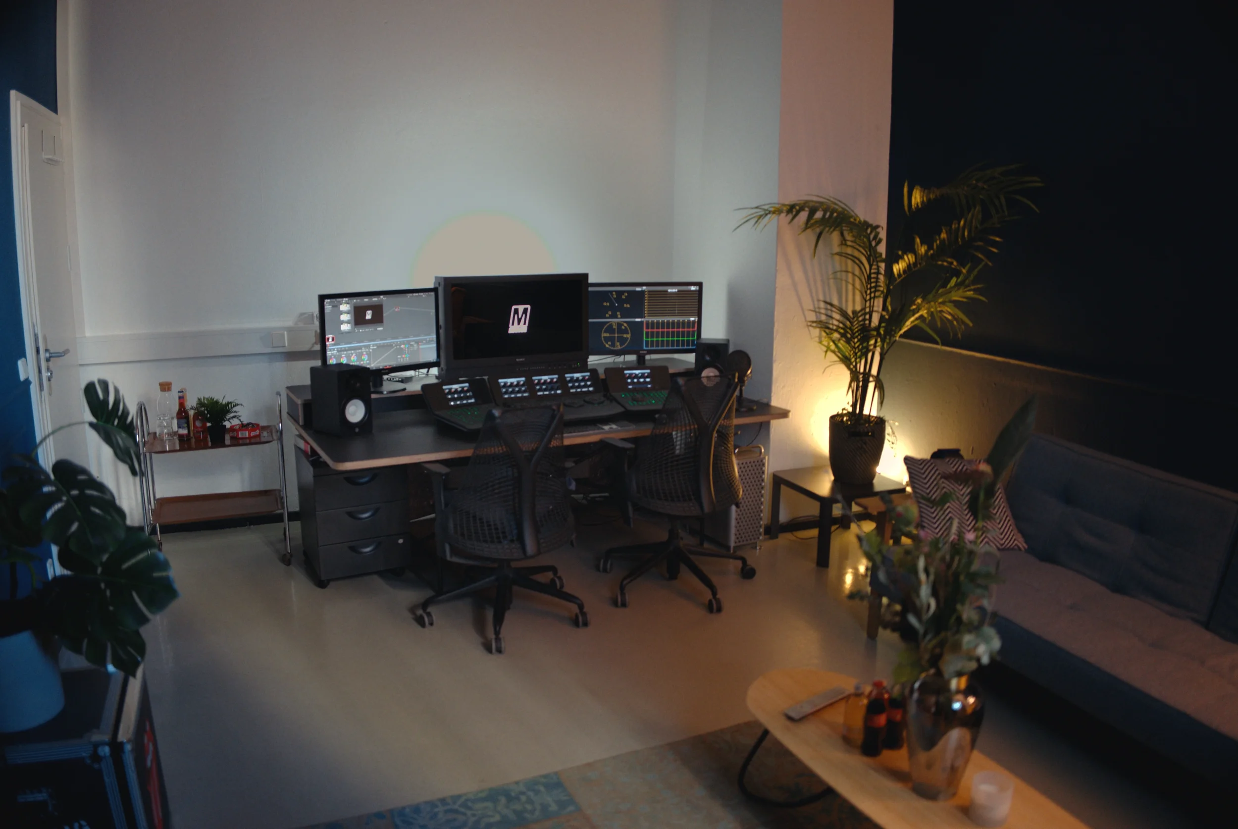 Wide view of the set up in Suite 1 or 2, furnished with a drinks station, a desk with multiple monitors, panels and speakers, a comfy couch and a coffee table.