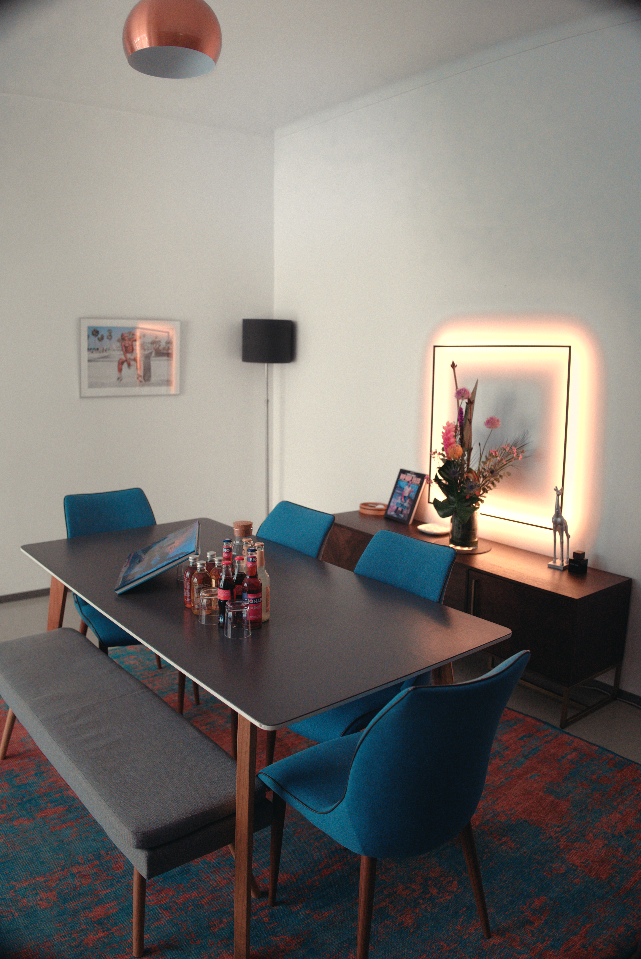 View of MOGRADE's studio space, furnished with a wood table, blue upholstered chairs, framed art and a neon glow wall art in the background.