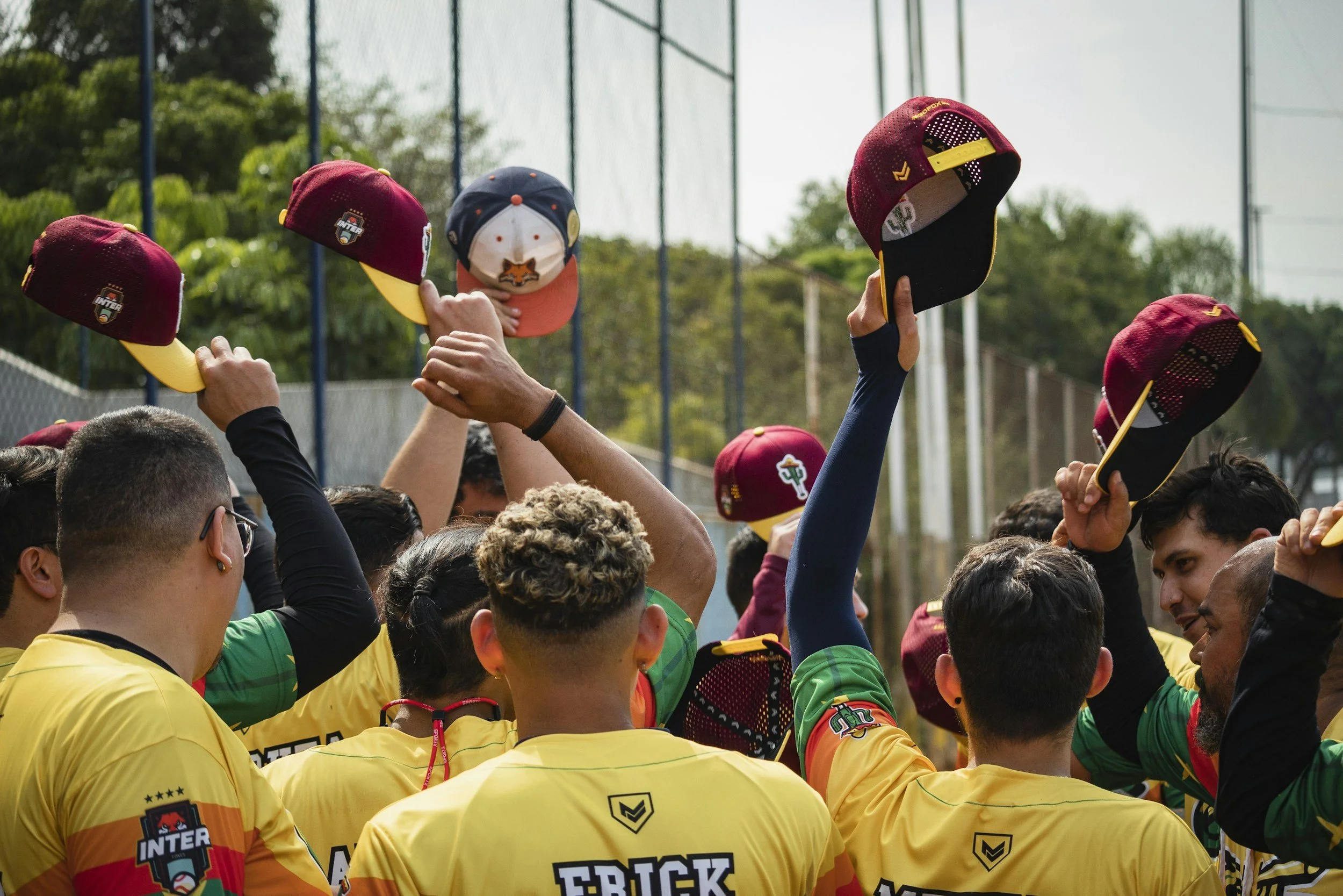 A group of baseball players wearing yellow jerseys and maroon caps lifting their caps into the air in a celebratory gesture on an outdoor field.