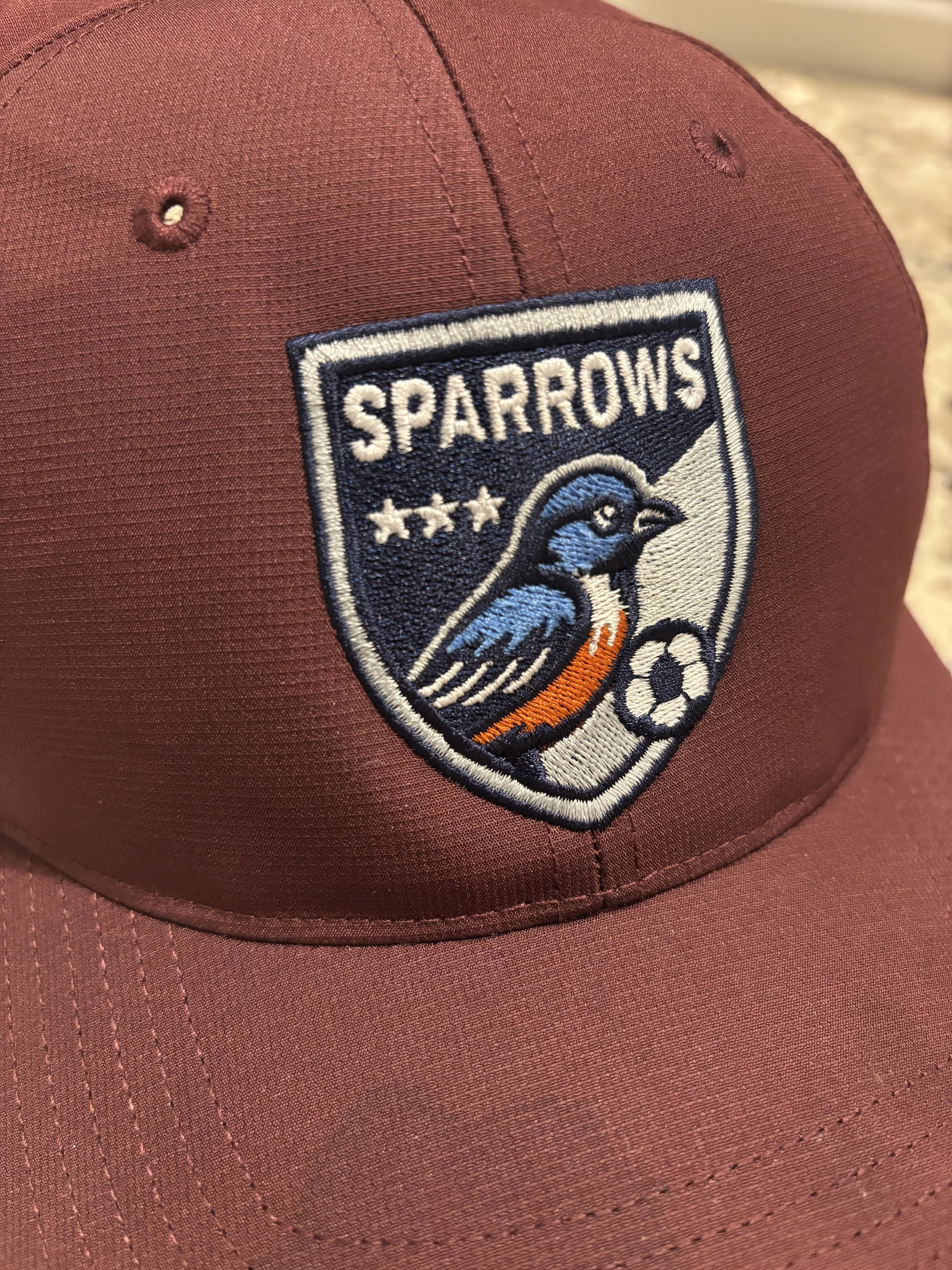 A maroon baseball cap with an embroidered patch of the Sparrows sports team logo, featuring a blue and orange bird, a soccer ball, and three stars above the bird, representing three seasons playing together.