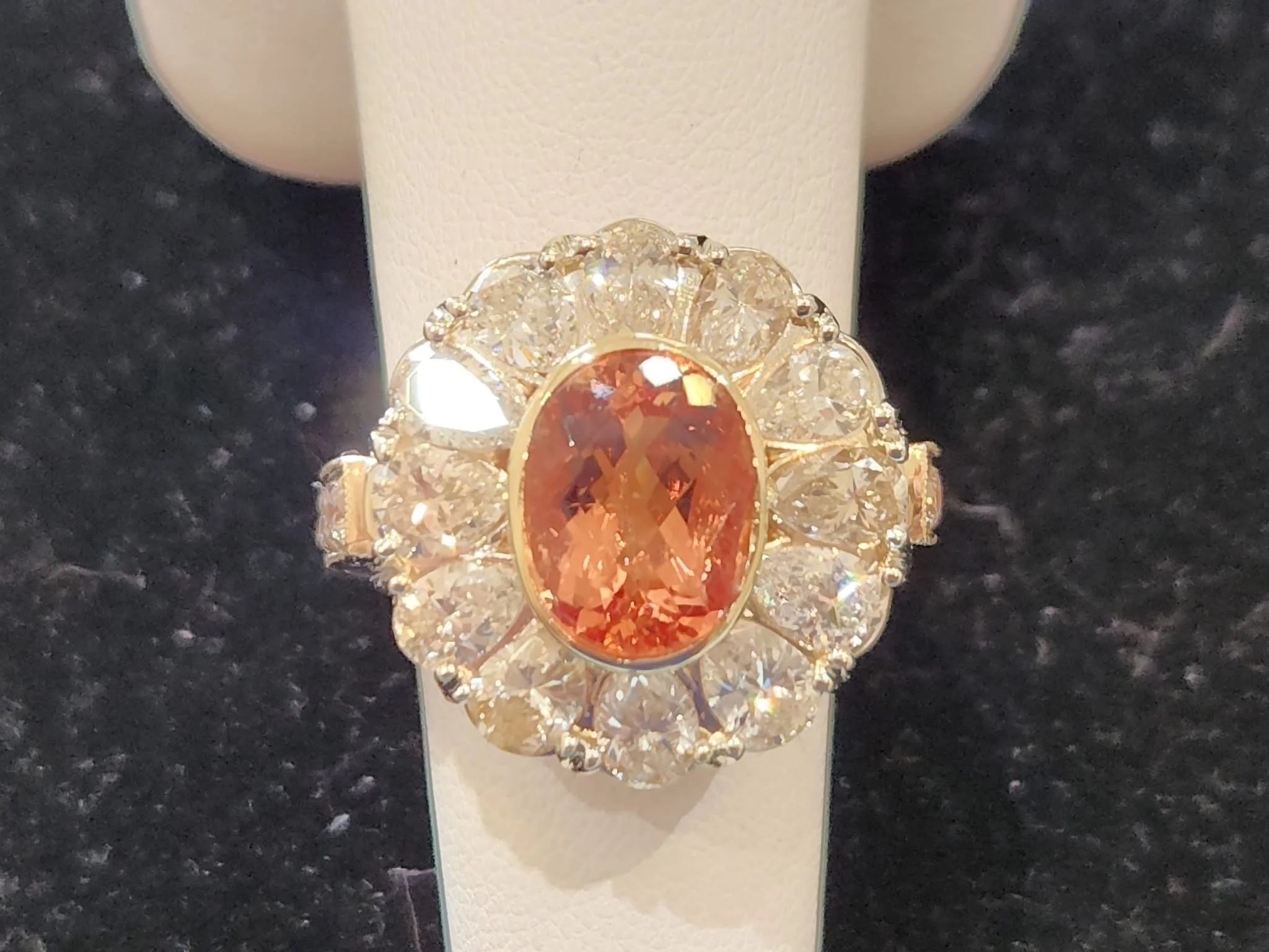 Gold ring with a large orange gemstone in the center, surrounded by smaller clear diamonds, on a white display stand.