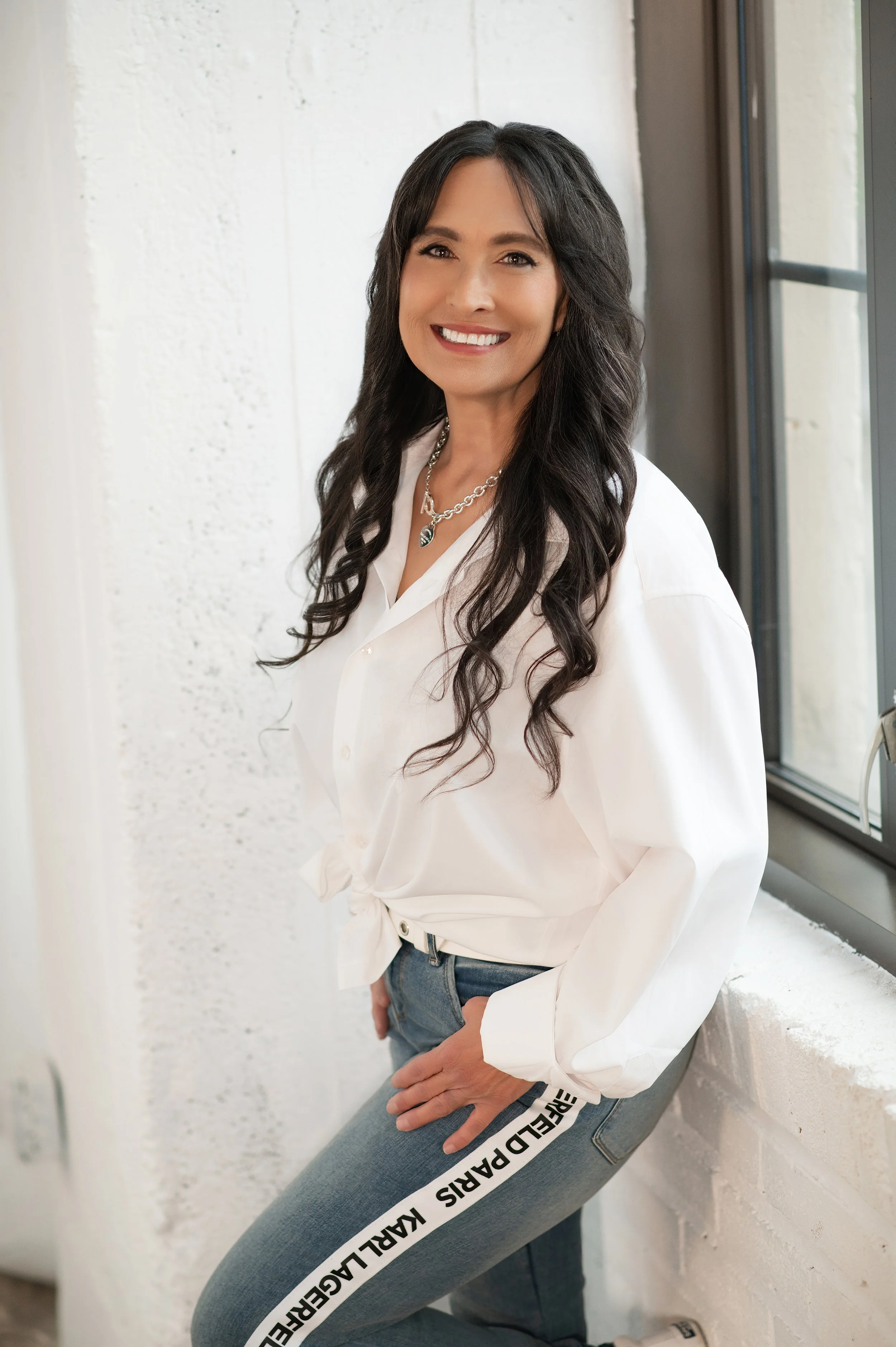 A woman with long dark curly hair, smiling, wearing a white shirt with knotted front, blue jeans with black and white text on the side, and layered necklaces, standing beside a window.