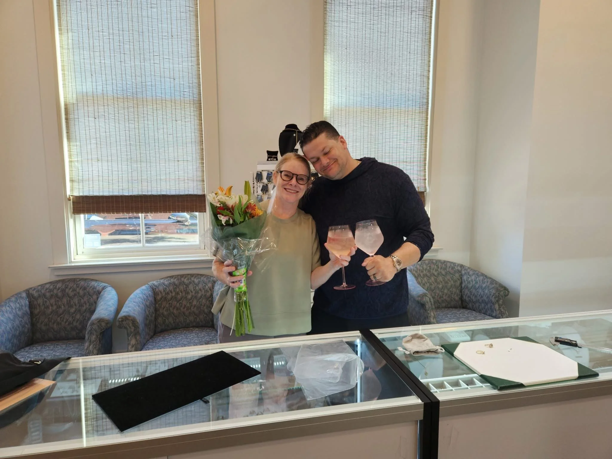 A woman and a man celebrating indoors, holding glasses of pink drinks; the woman holds a bouquet of flowers, and both are smiling, standing in front of a display case with jewelry.