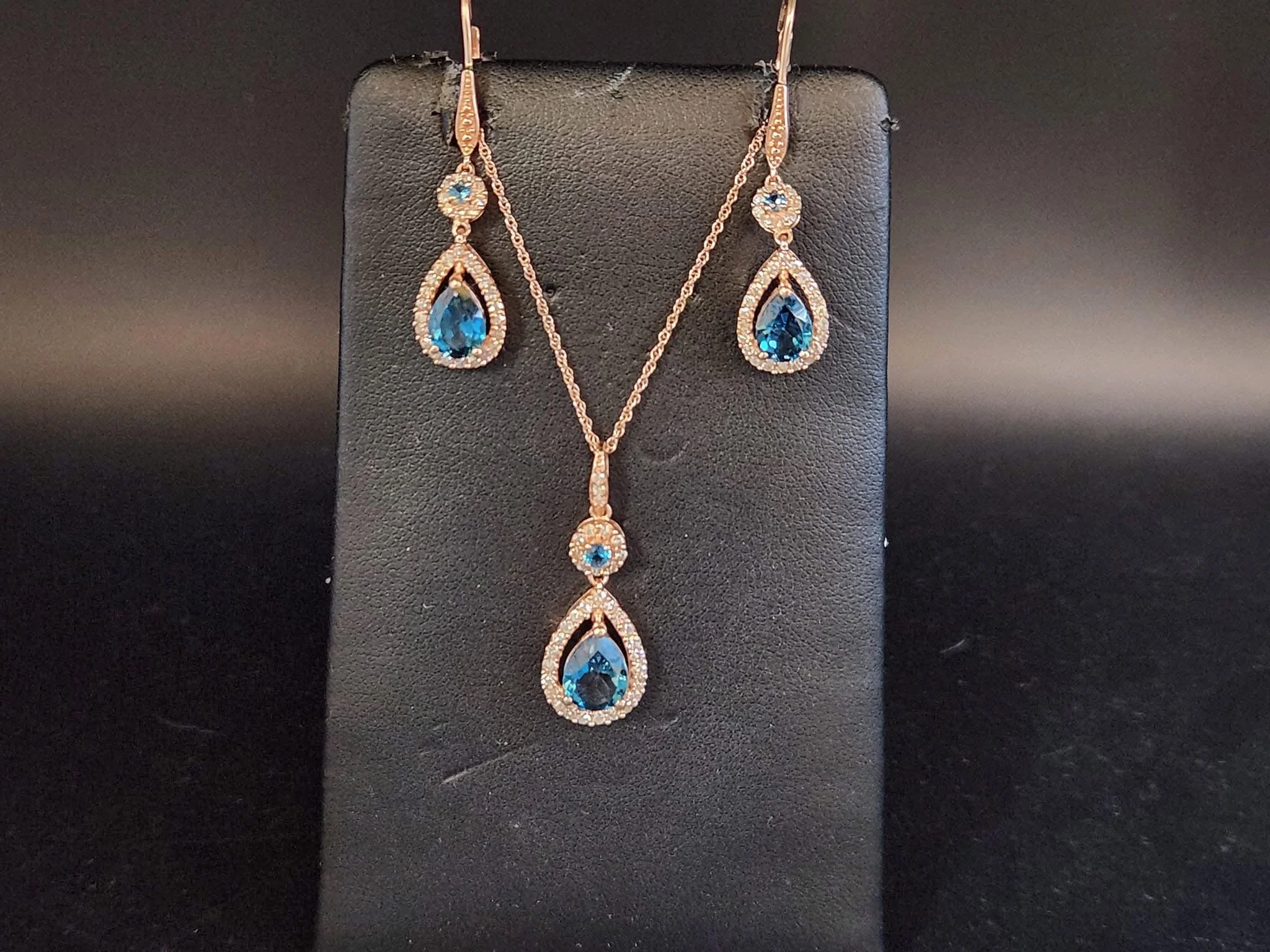 A jewelry set displayed on a black stand, including a pair of earrings and a matching necklace, each featuring a large pear-shaped blue gemstone surrounded by smaller clear stones.