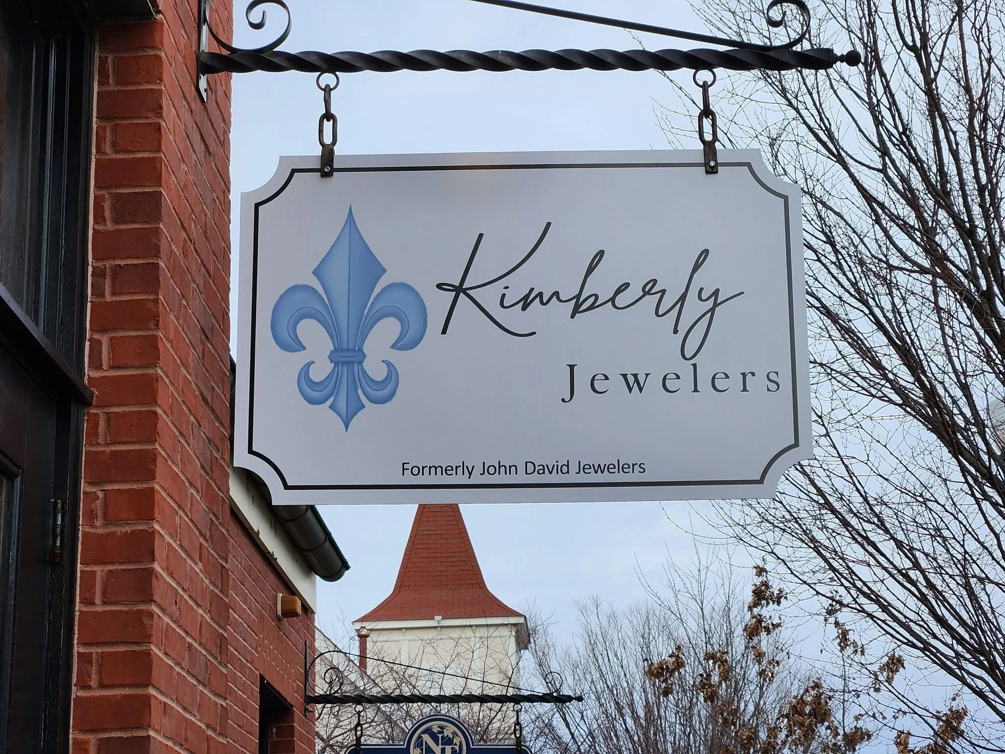 Business sign for Kimberly Jewelers with a blue fleur-de-lis logo, on a brick building with a church steeple visible in the background and leafless trees.