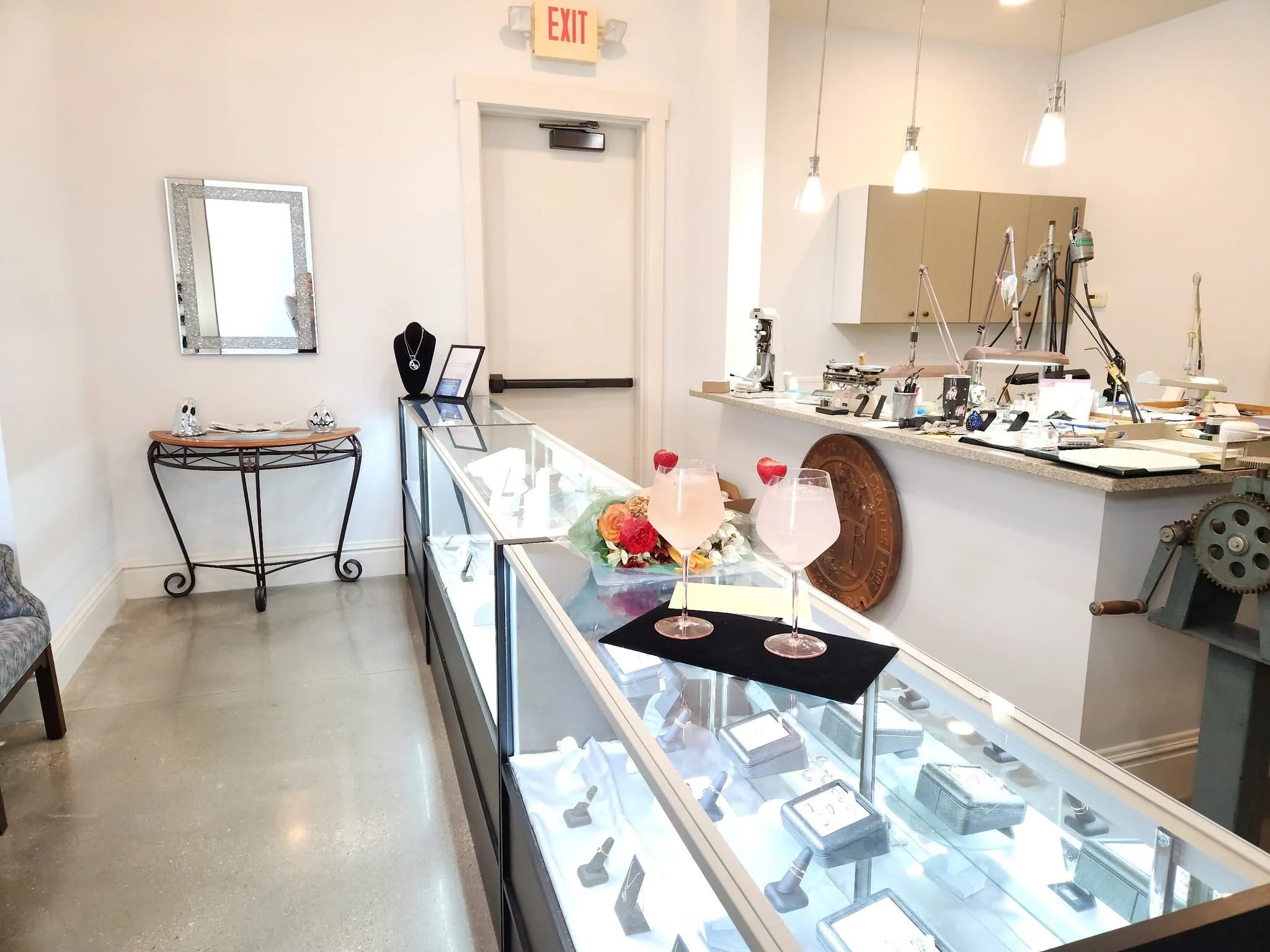 Jewelry store interior with a glass display case filled with jewelry, a jewelry necklace on a black bust, a small table with jewelry items, a decorative mirror on the wall, a counter with various tools, and two pink drinks with flowers on a black napkin.