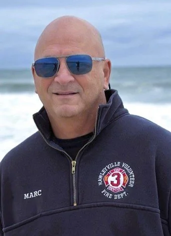 Man wearing sunglasses and a navy zip-up jacket with 'MARC' and 'Vanderbilt Volunteer Fire Department' logo, standing on the beach with ocean waves in the background.