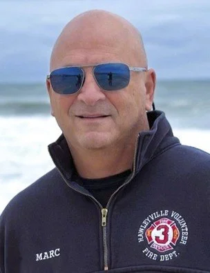 A bald man wearing sunglasses and a navy jacket standing on a beach with the ocean in the background.