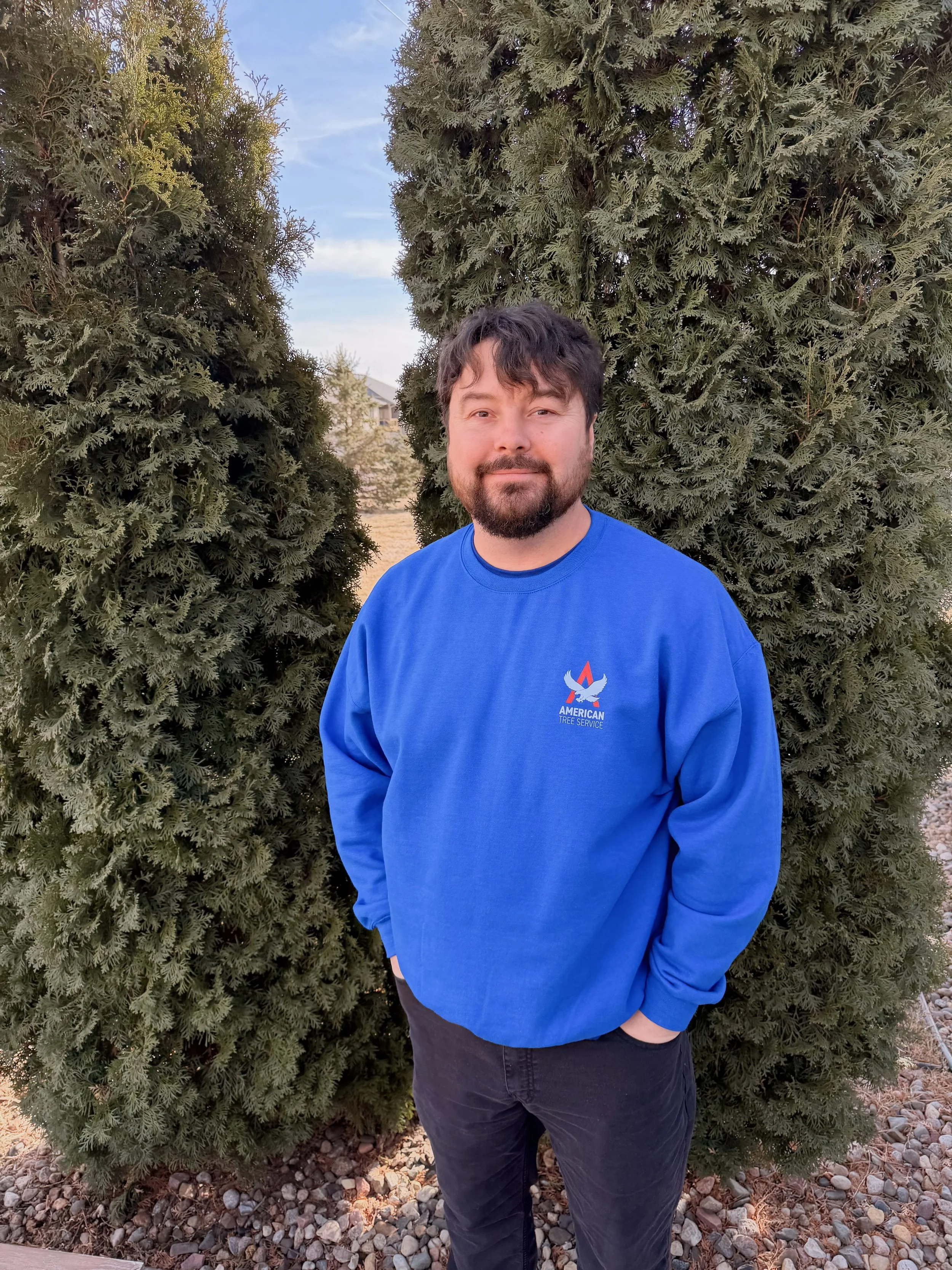 A man with dark hair and a beard is standing outdoors in front of two tall evergreen trees. He is wearing a blue sweatshirt with the logo of American Tree Service and has a slight smile. The background shows a clear sky and some distant trees.