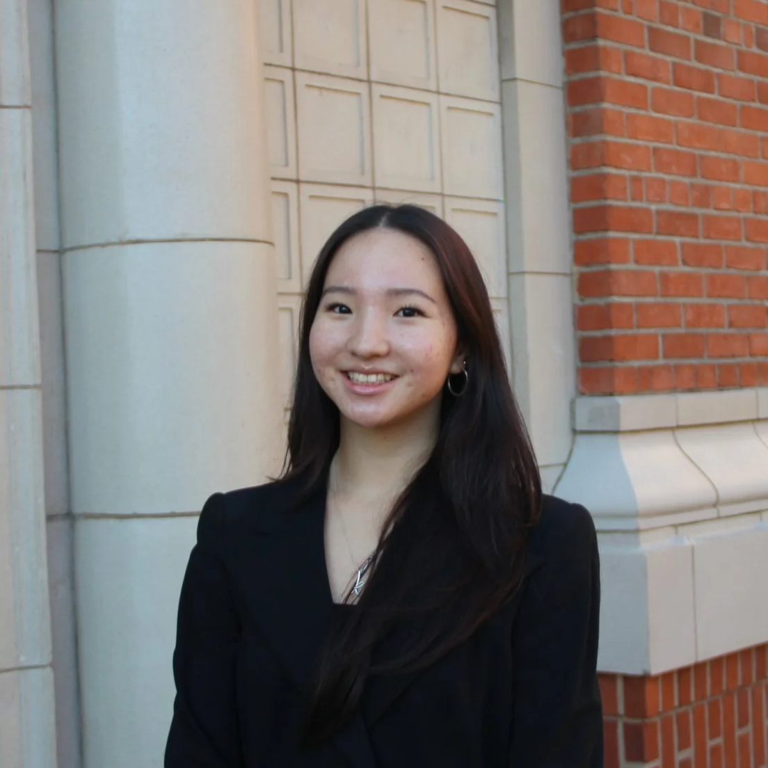 Young woman with long dark hair smiling, wearing a black blazer, standing outdoors by a building with brick and light-colored stonework.