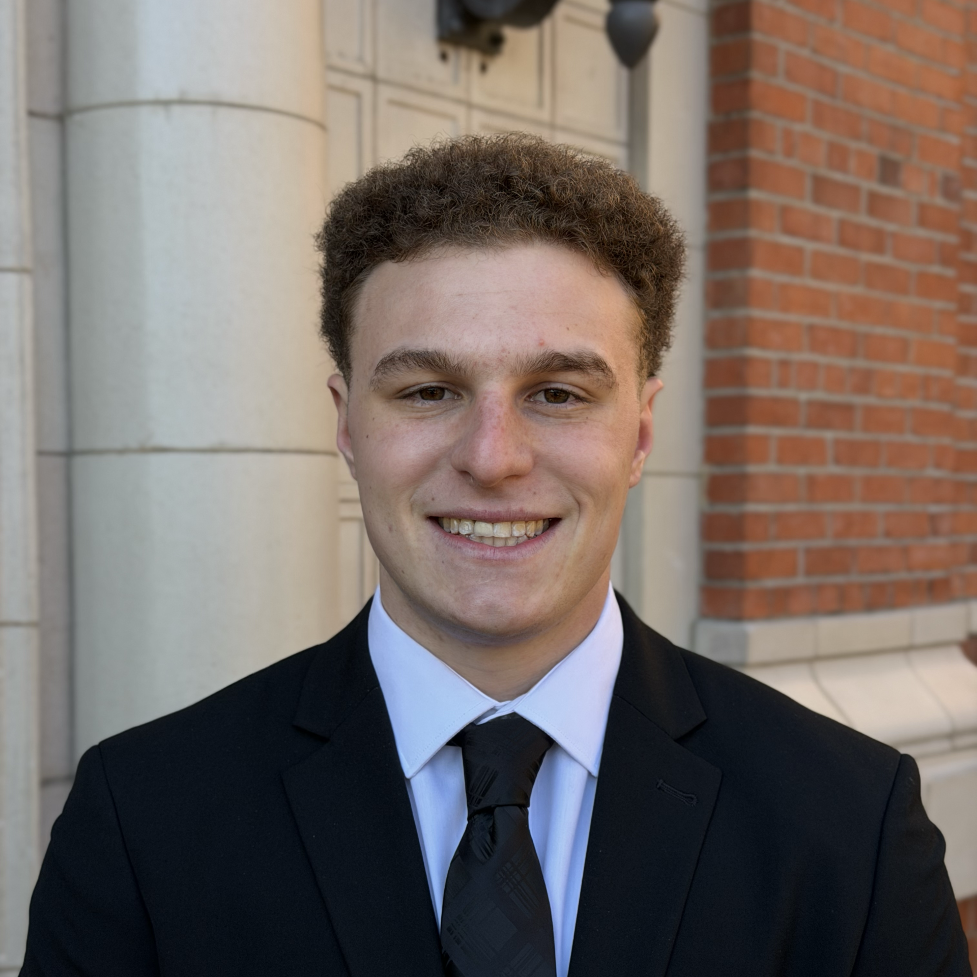 Portrait of a young man in a black suit and tie, smiling outdoors in front of a brick and stone building.
