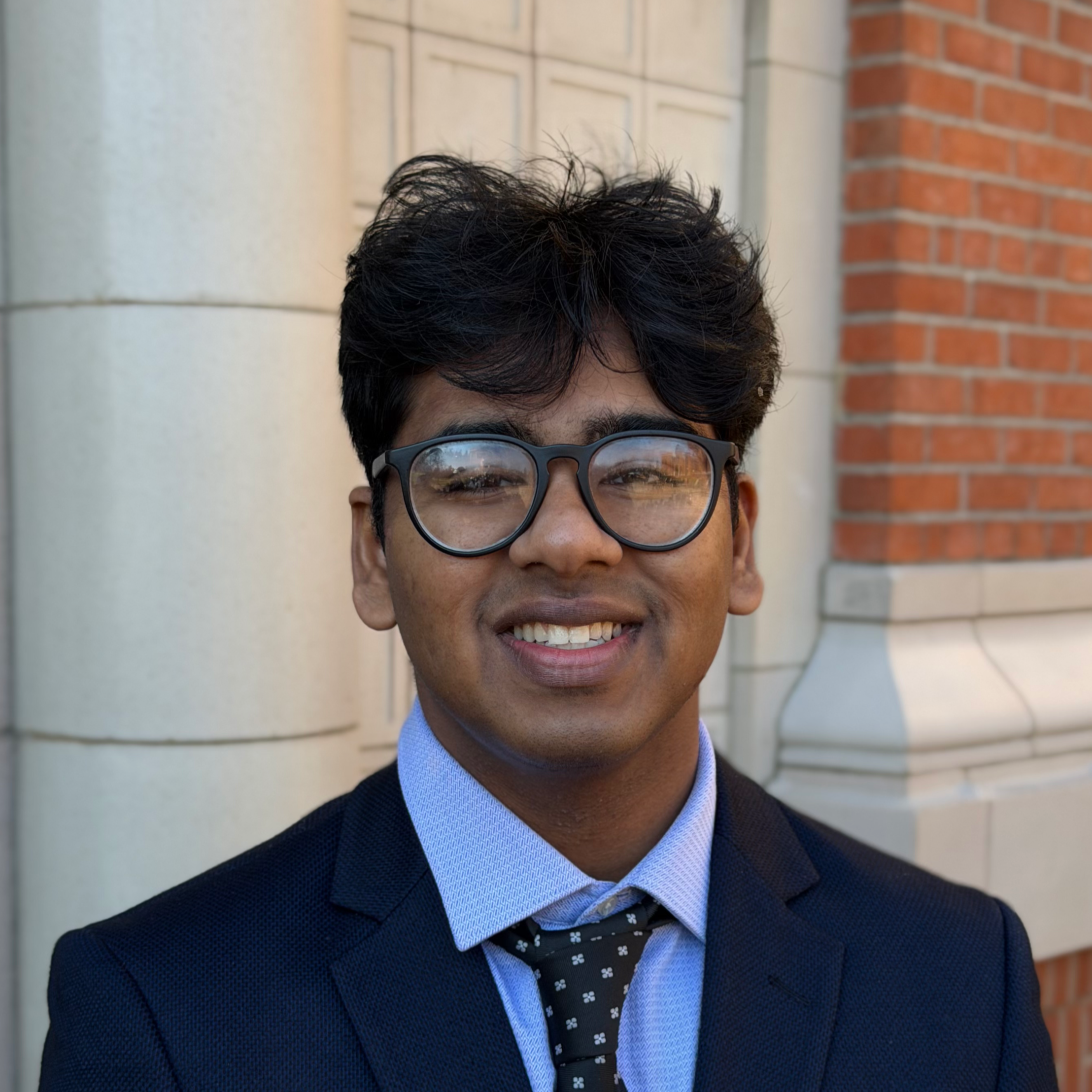 Young man in suit and glasses smiling outdoors in front of modern office building with a large yellow letter 'D' sign.