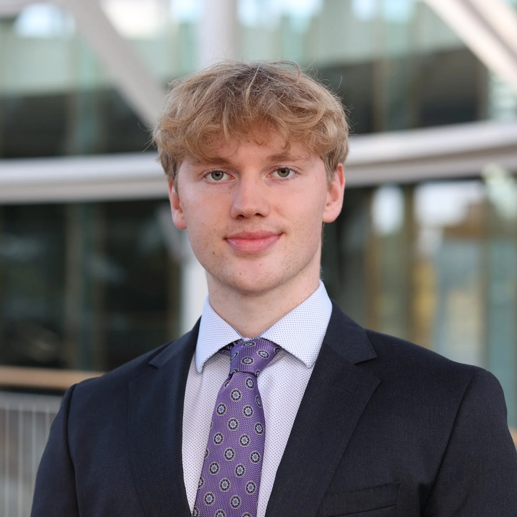 Portrait of a young man with blonde hair wearing a navy suit, white shirt, and purple patterned tie, standing outdoors in front of a modern glass building.
