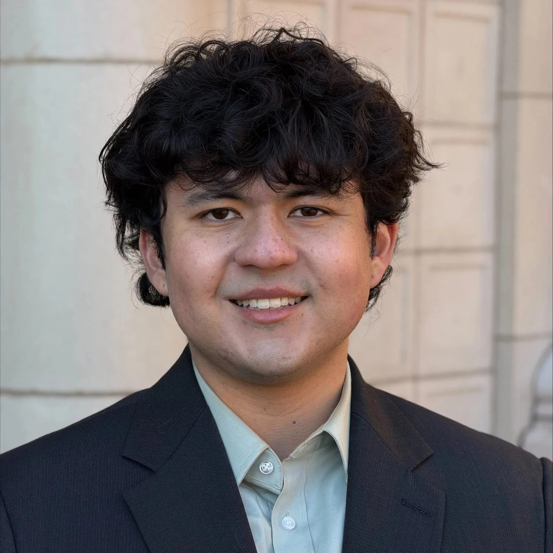 A young man with medium-length, curly black hair and light skin, wearing a dark suit jacket and light-colored dress shirt, smiling in front of a beige brick building.